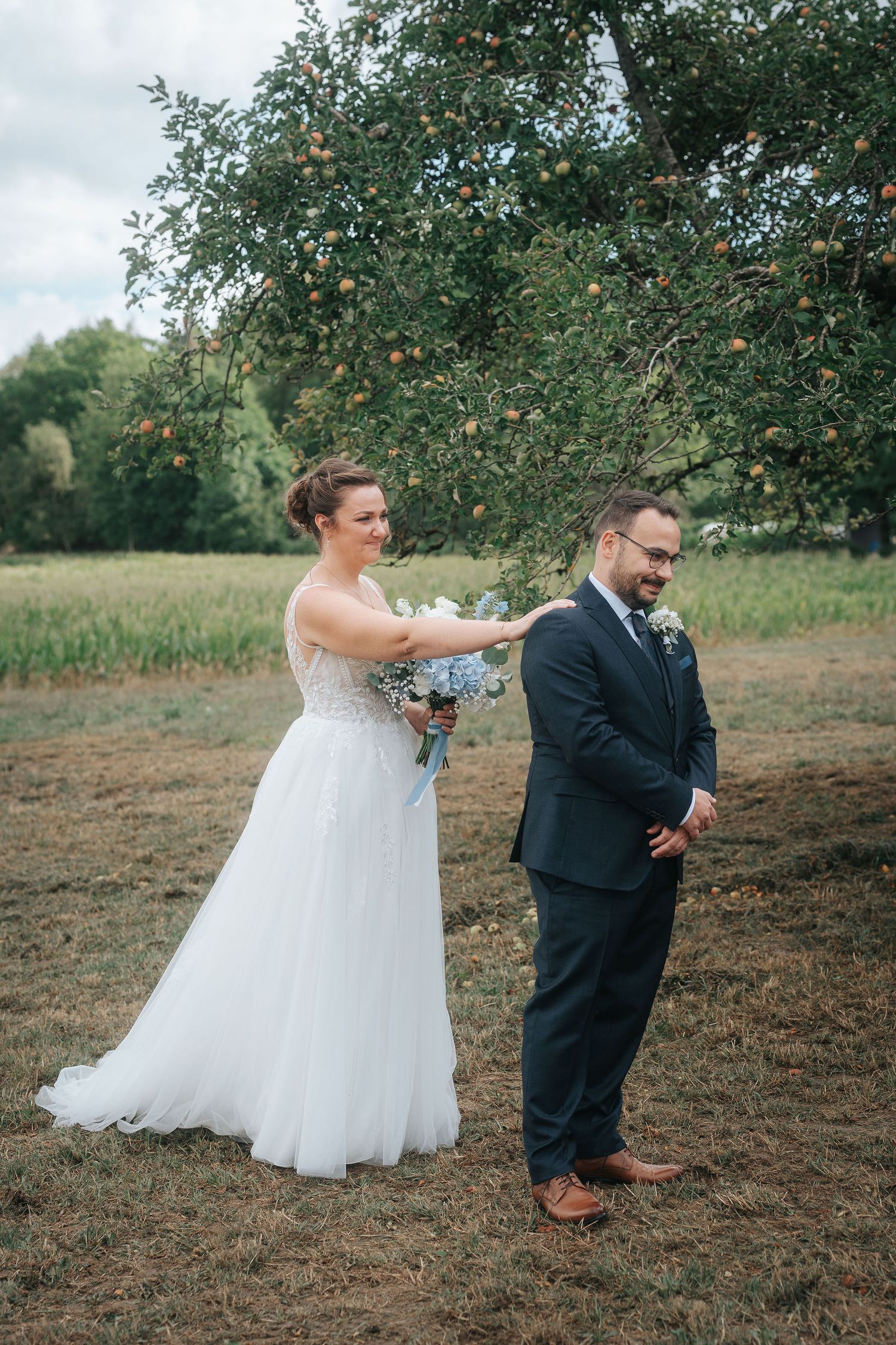Bride in a white tulle gown holding a blue bouquet reaches out to tap the groom, who stands facing away, under an apple tree.