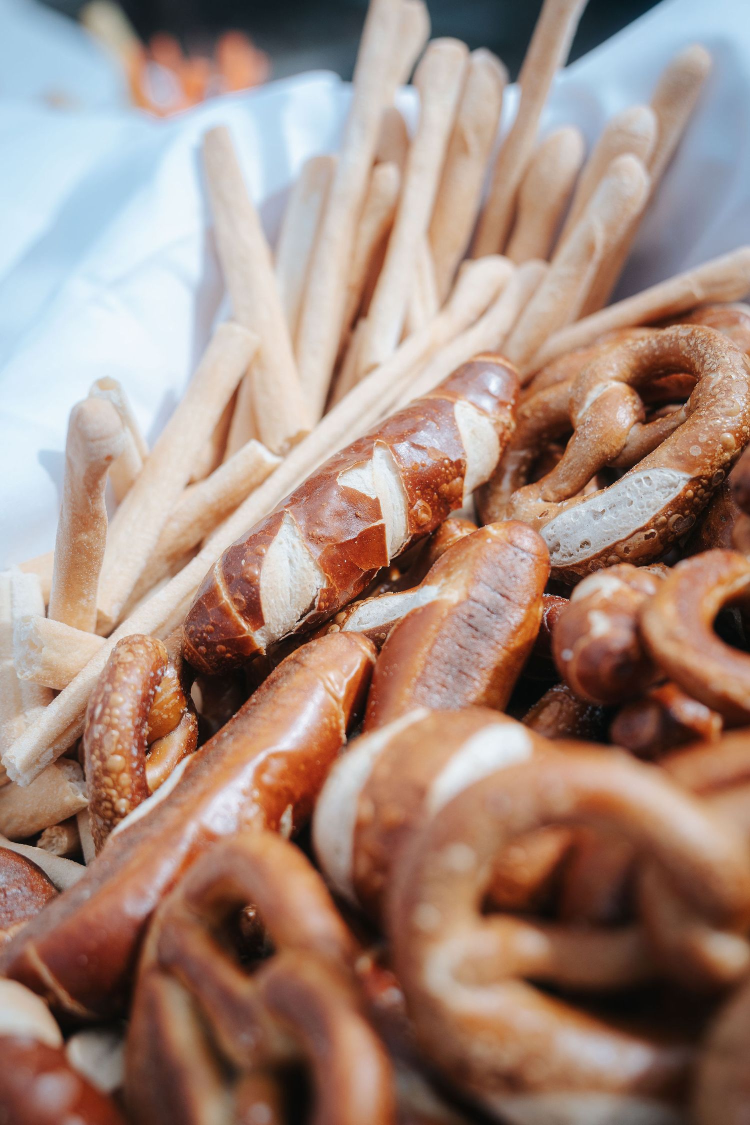 Freshly baked soft pretzels with salt and crunchy breadsticks piled together on a white cloth at a bakery or buffet display.