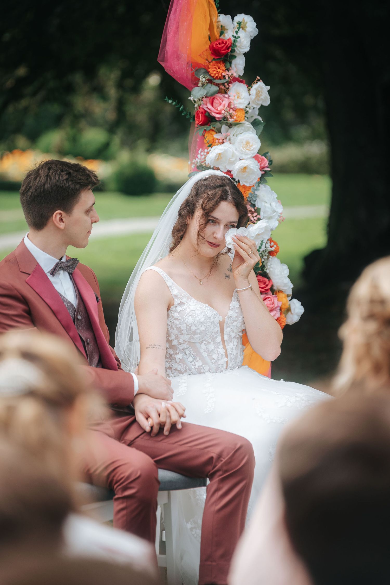 Emotional bride in a lace gown and veil dabs her tears with a tissue beside her groom in a rust suit at an outdoor wedding ceremony.