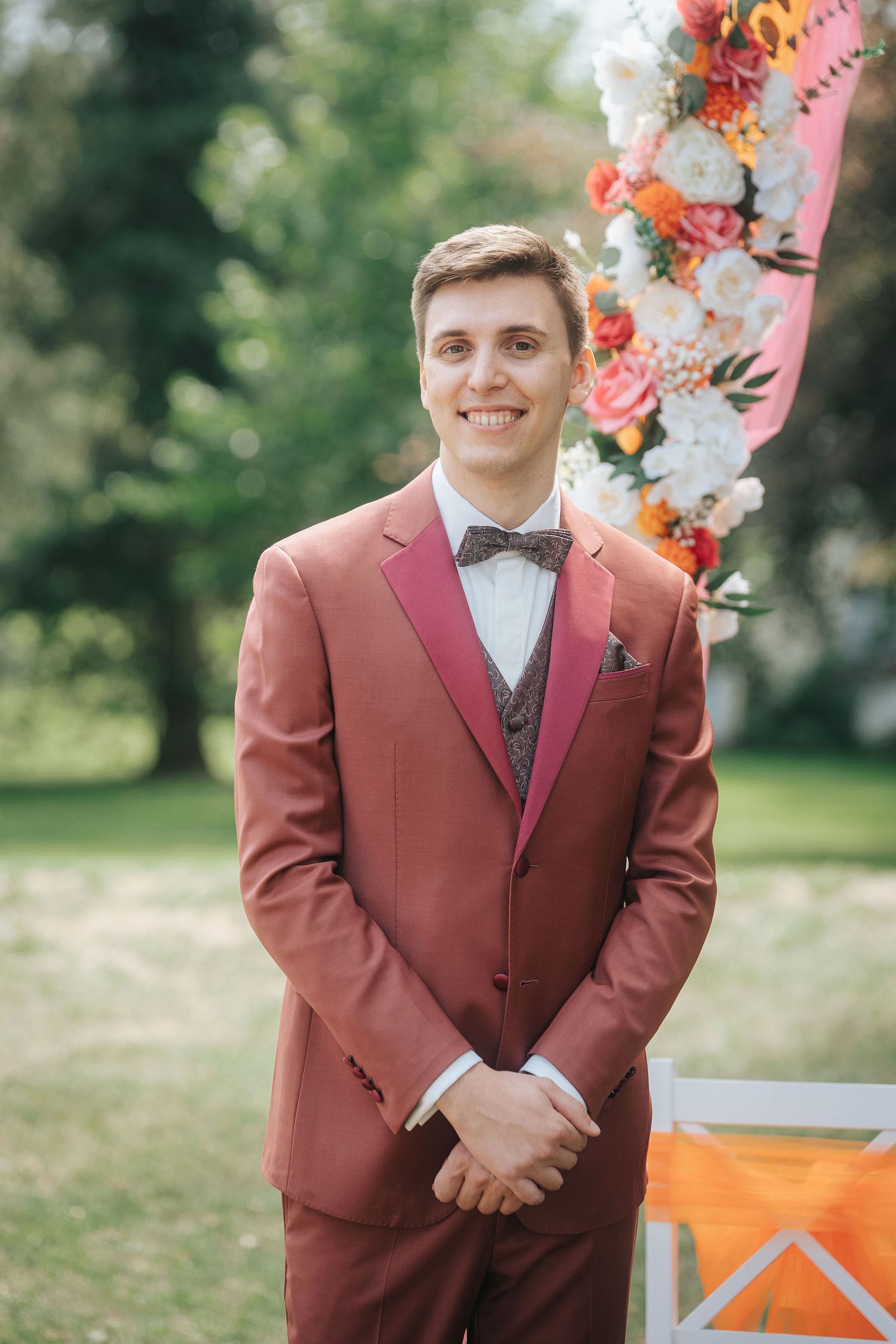 Smiling groom in a rust-colored suit and brown bow tie stands outdoors beside a floral wedding arch with pink, white and orange blooms.