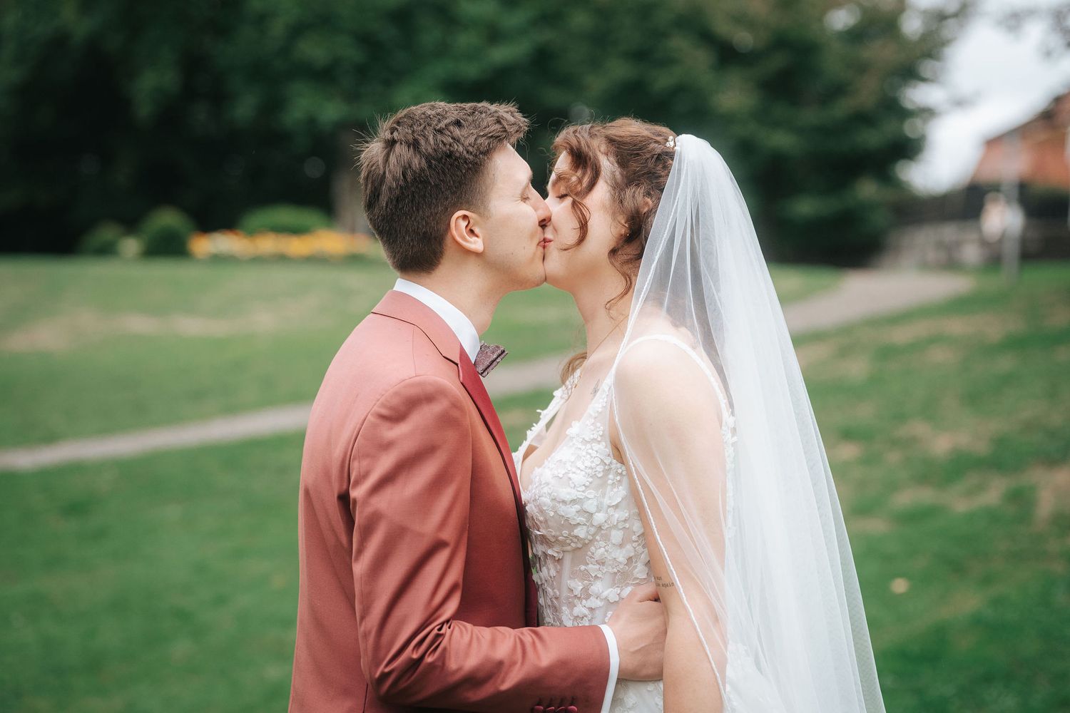 Bride in a lace gown and long veil kisses groom in a rust-colored suit on a green lawn during their outdoor wedding.