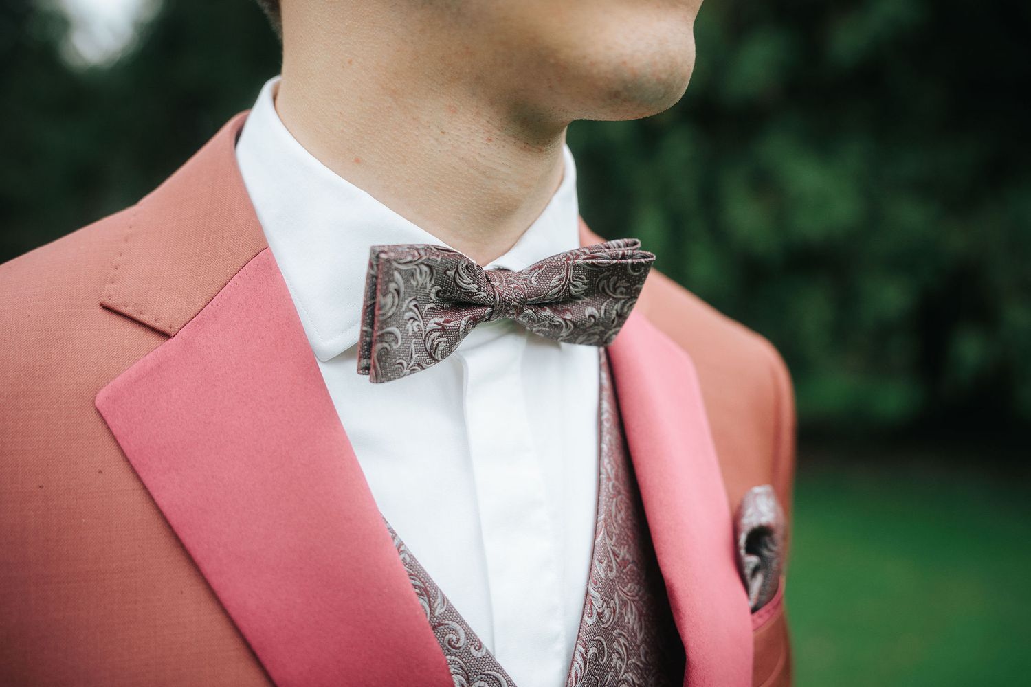 Man in a coral pink suit jacket and white shirt wearing a paisley bow tie with matching pocket square outdoors.