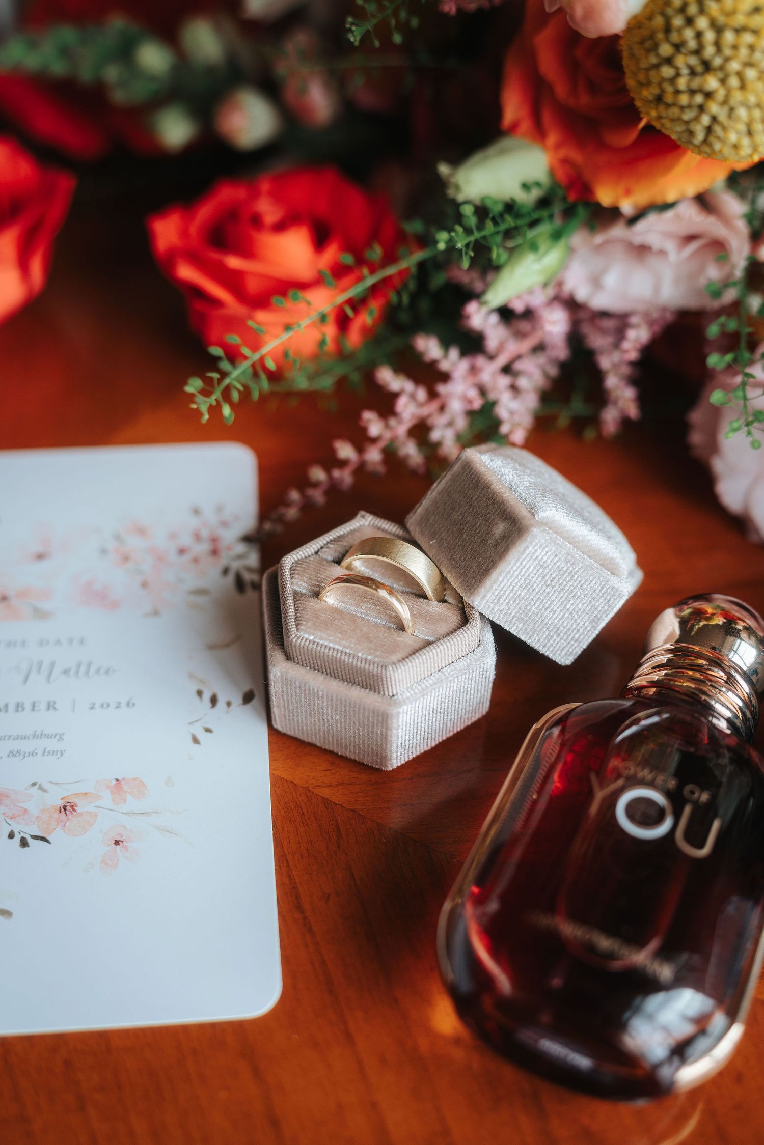 Gold wedding bands in a velvet ring box beside a perfume bottle, floral bouquet and invitation card on a wooden table.