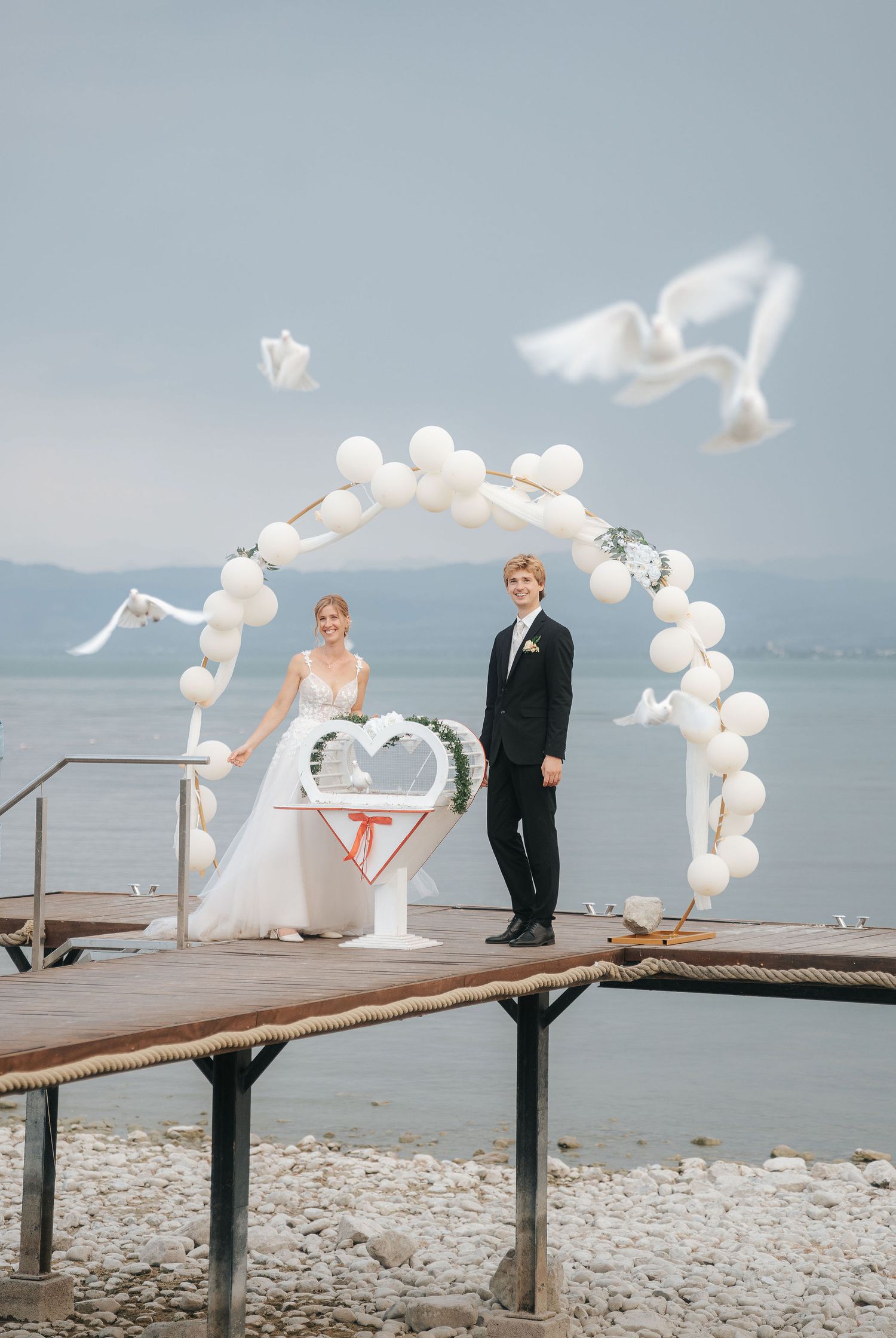 Bride and groom release white doves under balloon arch on lakeside pier during outdoor wedding ceremony.