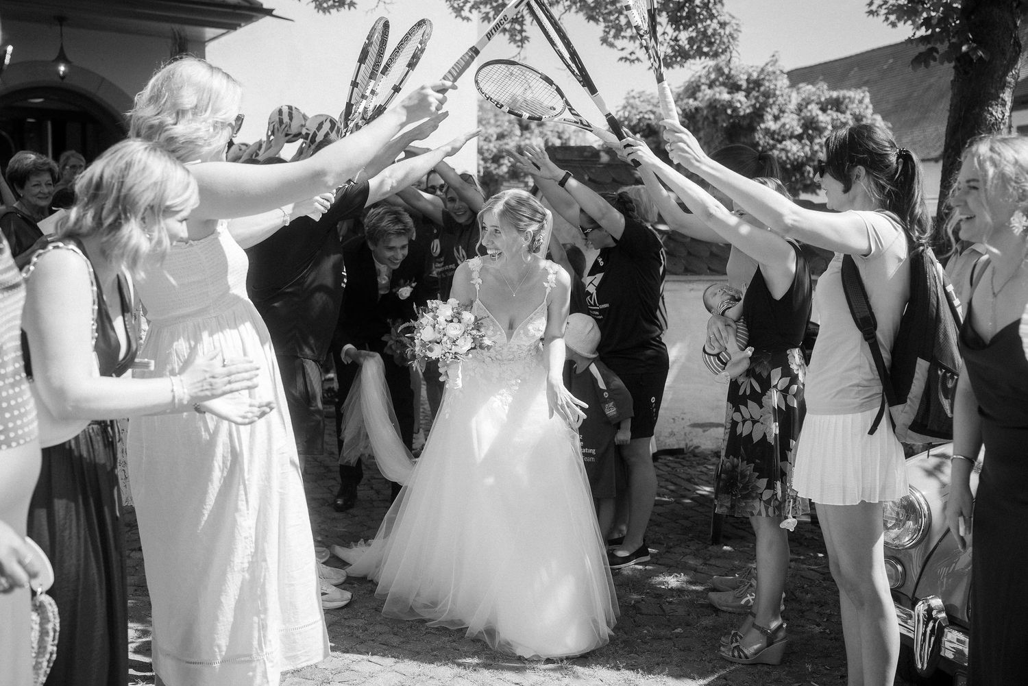 Black and white photo of a bride walking through an arch of raised tennis rackets held by wedding guests outdoors.