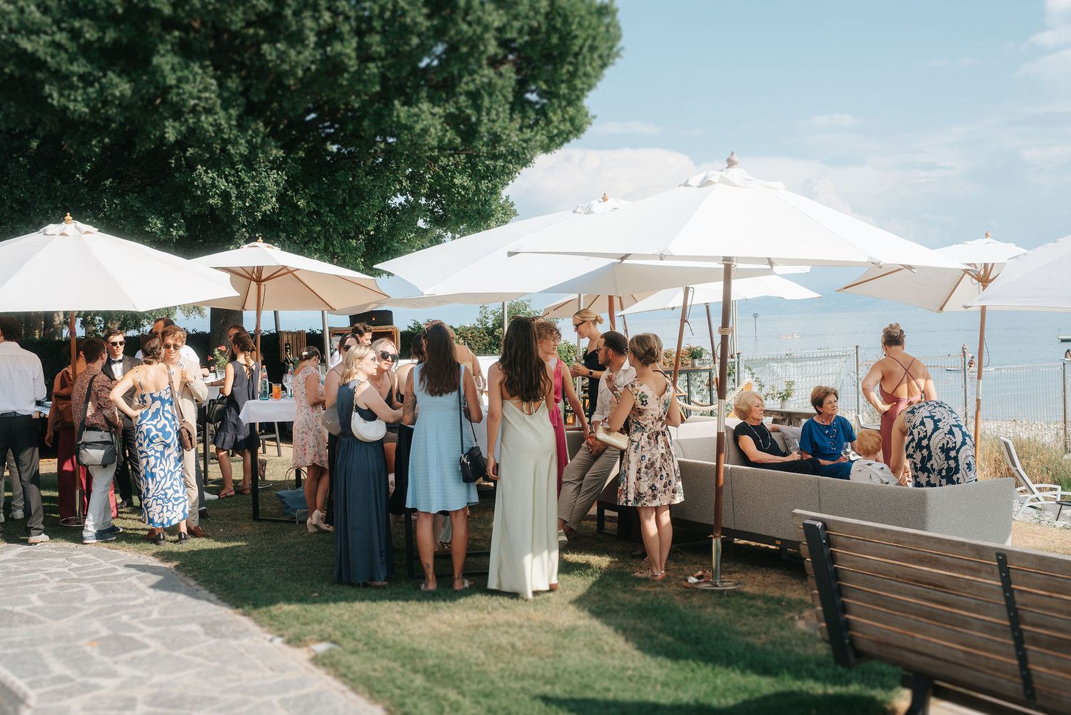 Guests mingle outdoors under white umbrellas at an elegant garden wedding reception on a sunny day.