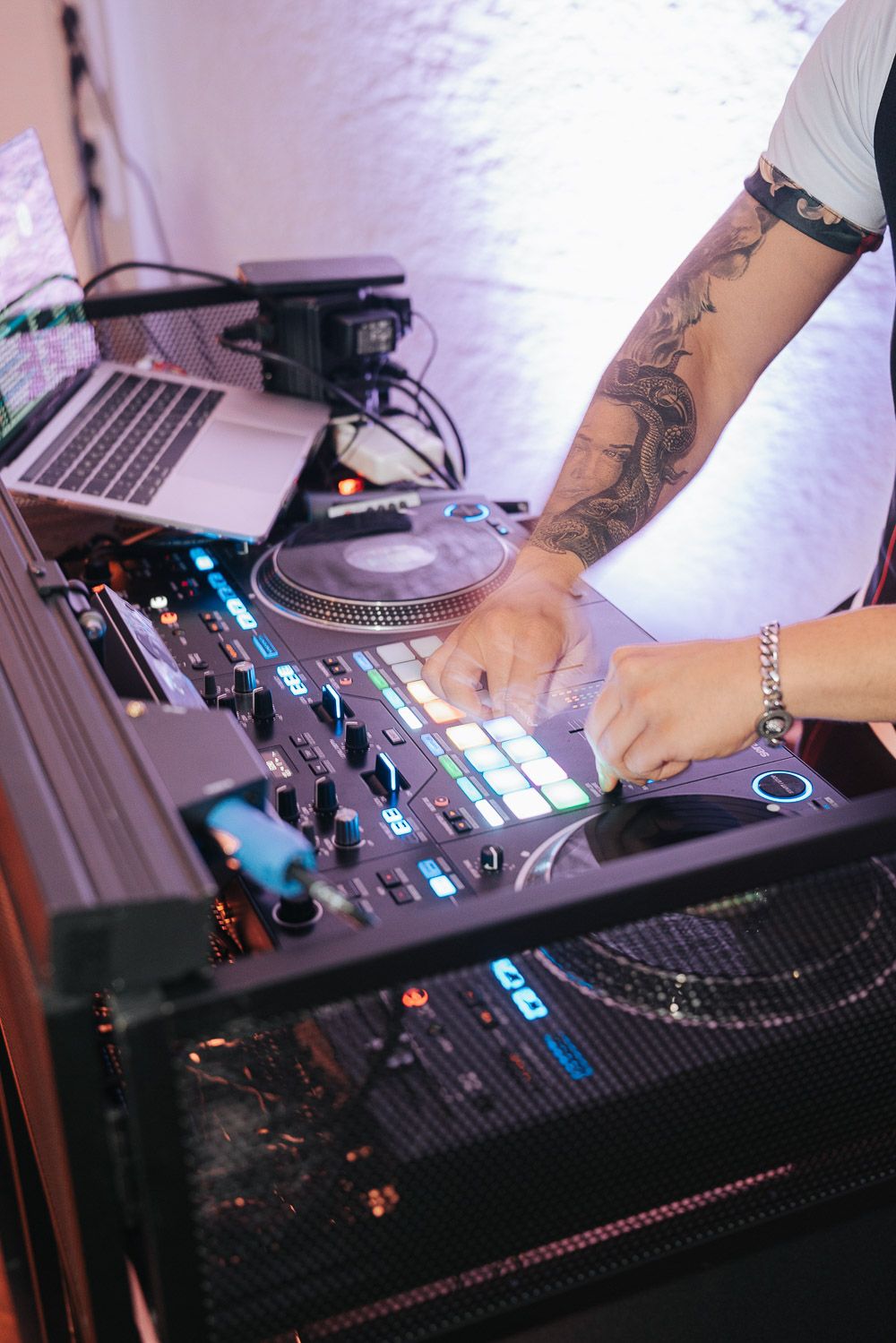 A DJ works their equipment with tattooed arms, adjusting controls on a mixing board with turntables and a laptop visible.
