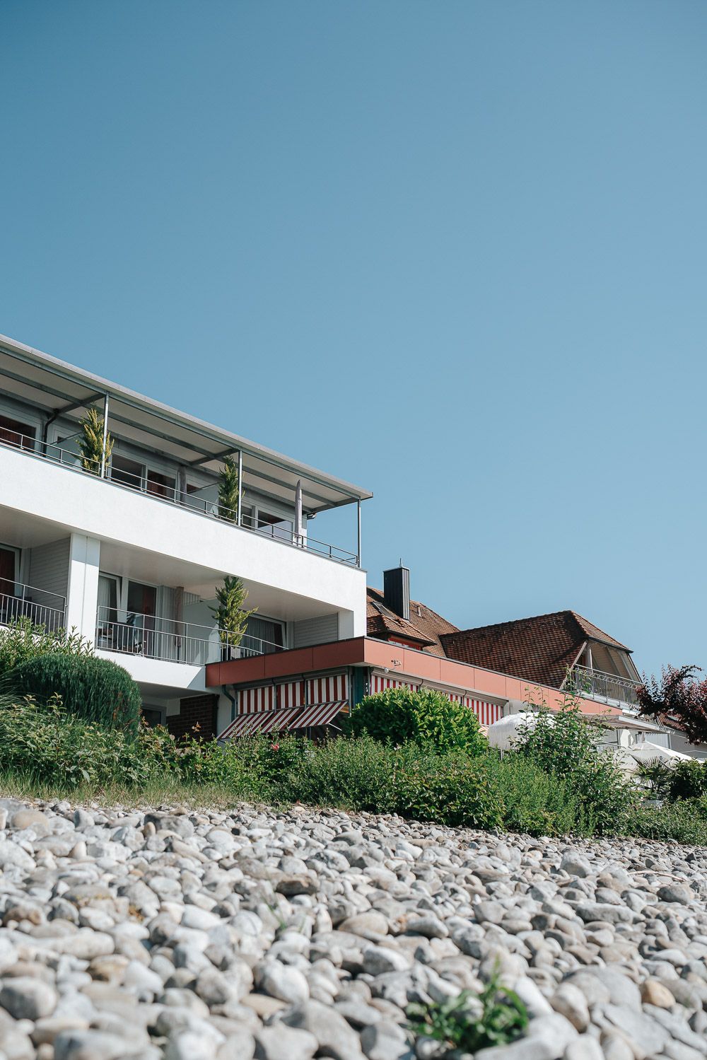 Modern hillside homes overlook a rock-covered shoreline against a clear blue sky.