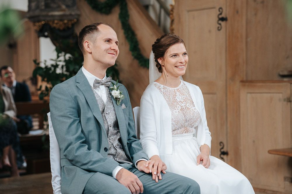 Two people sitting together at a rustic wedding ceremony, wearing a grey suit and elegant white lace dress.