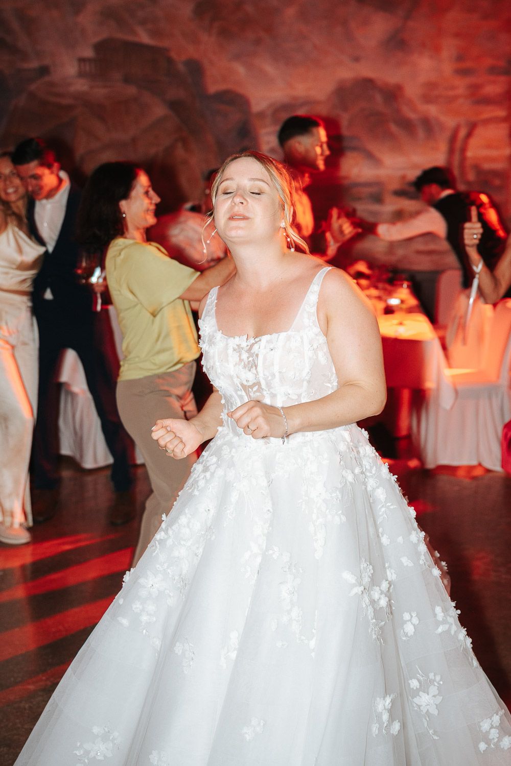 A bride in a white lace ballgown dances with joy on the reception dance floor under dramatic red lighting.