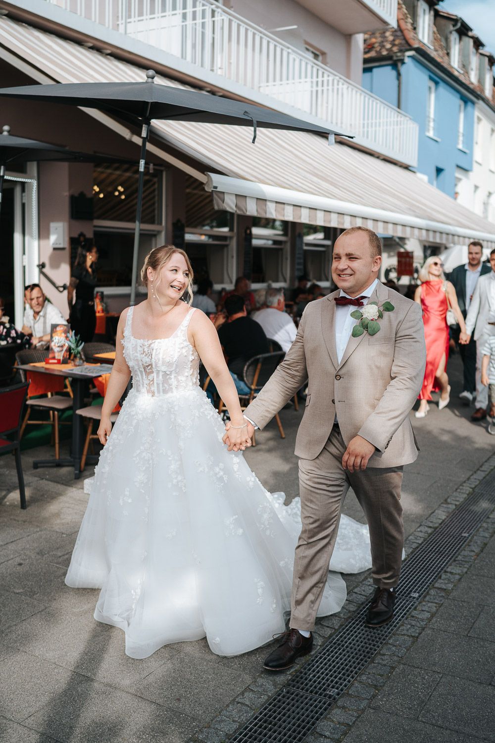 A couple in wedding attire walking hand-in-hand down a shaded storefront walkway with white columns.