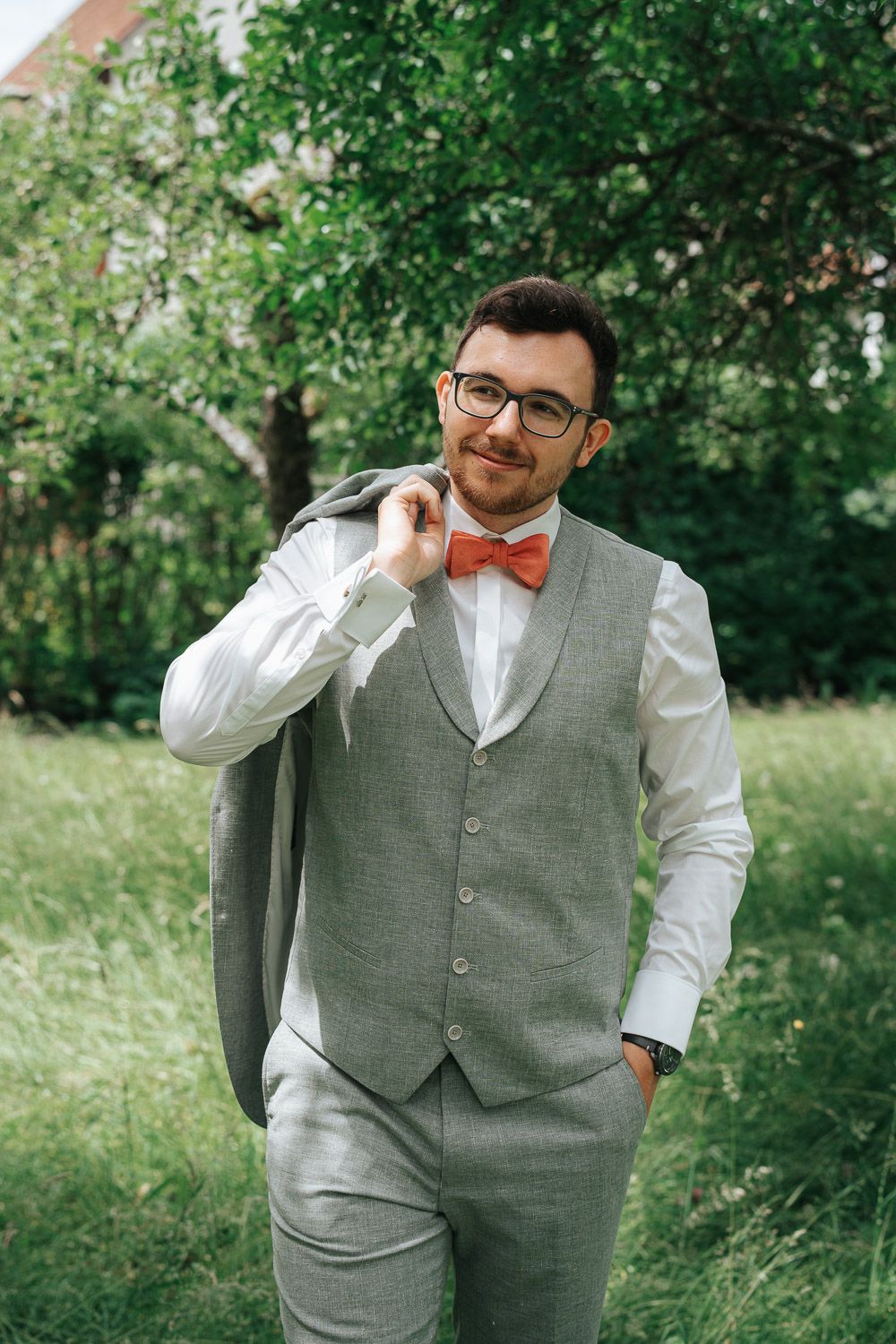 A person in a grey vest and bow tie poses outdoors in a natural garden setting.