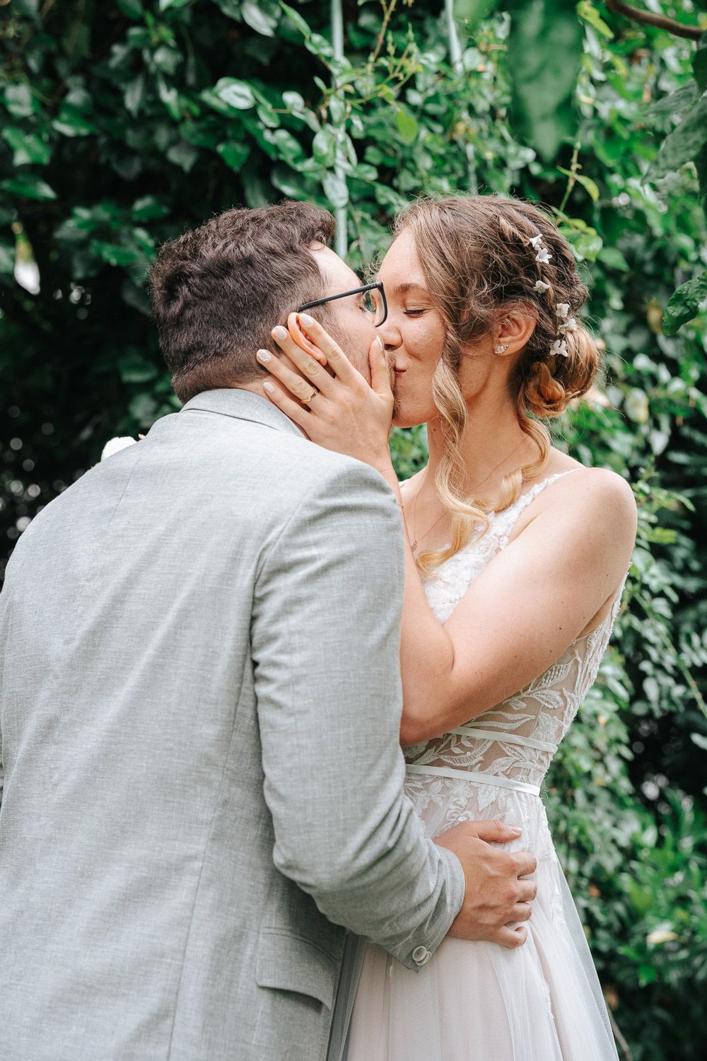 A romantic wedding couple sharing a tender kiss in a garden setting with lush green foliage in the background.