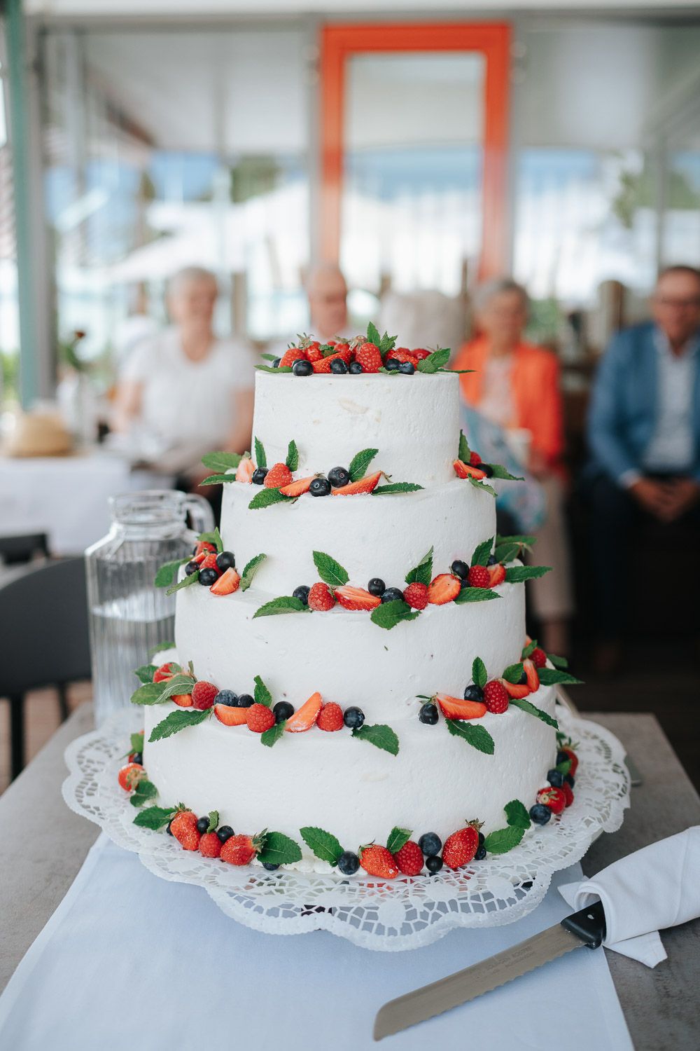 White three-tier wedding cake decorated with fresh strawberries, blueberries and green leaves on a lace doily.