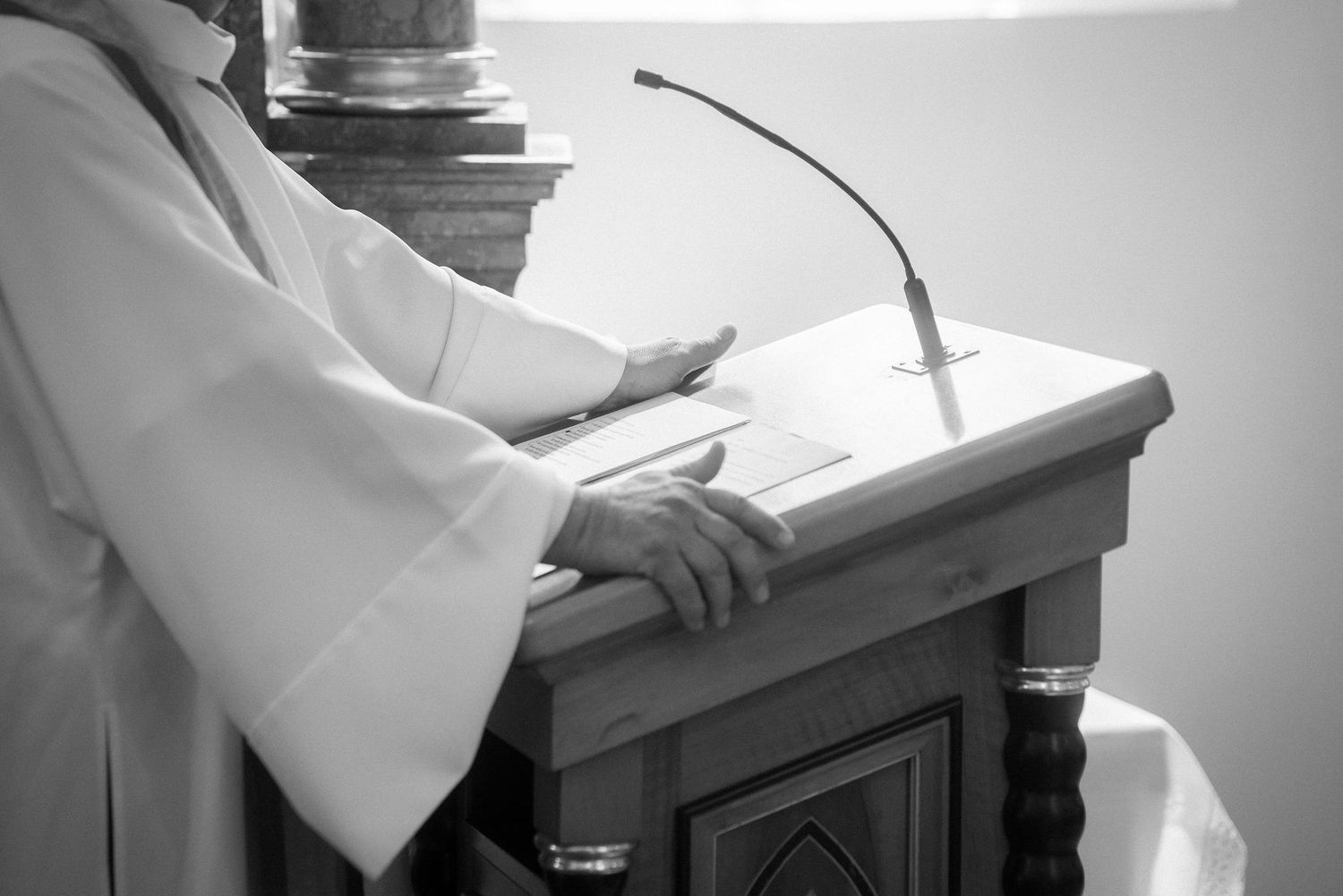 Black and white photo of a priest in white vestments resting his hands on a wooden church lectern with a microphone.