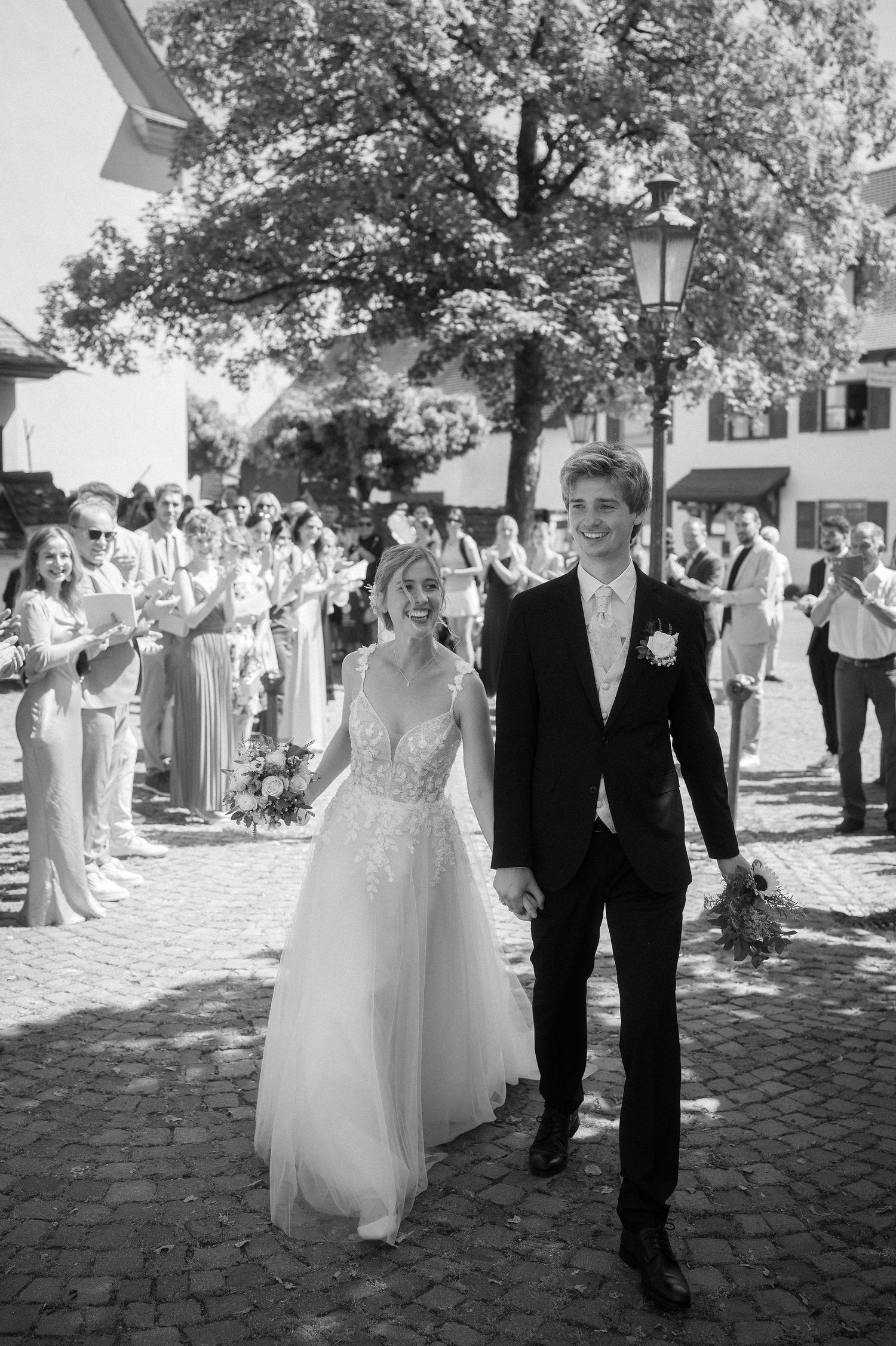 Smiling bride and groom walk hand in hand across a cobblestone square as cheering wedding guests applaud in this black and white photo.