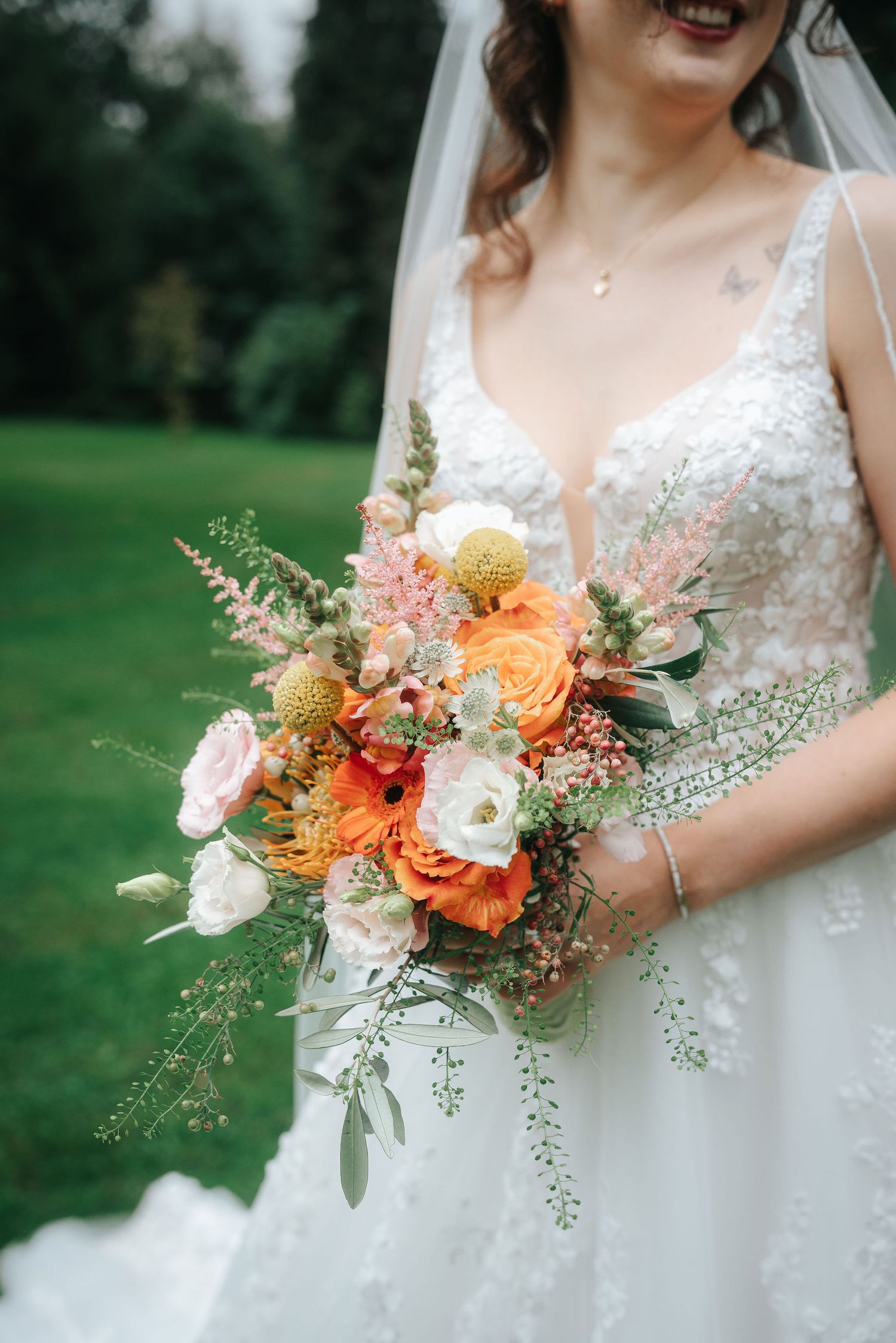 Bride in a lace wedding gown and veil holds a colorful bouquet of orange, cream and pink flowers with greenery on a lawn.