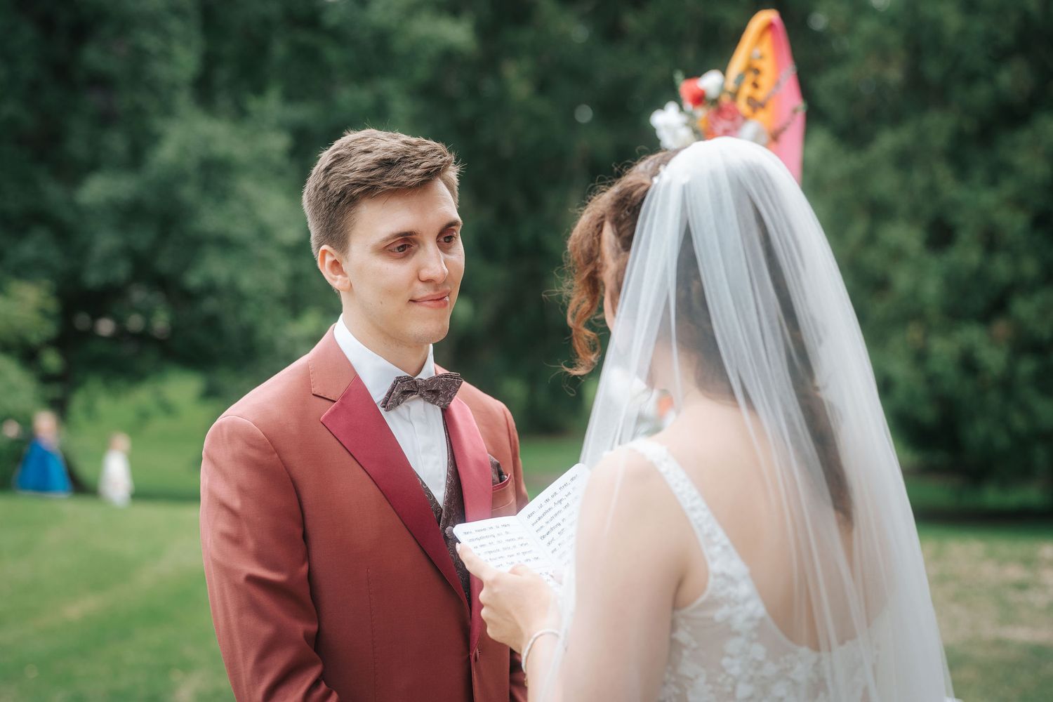 Groom in a rust-red suit and bow tie smiles at his veiled bride as she reads her vows during an outdoor wedding ceremony in a green park.