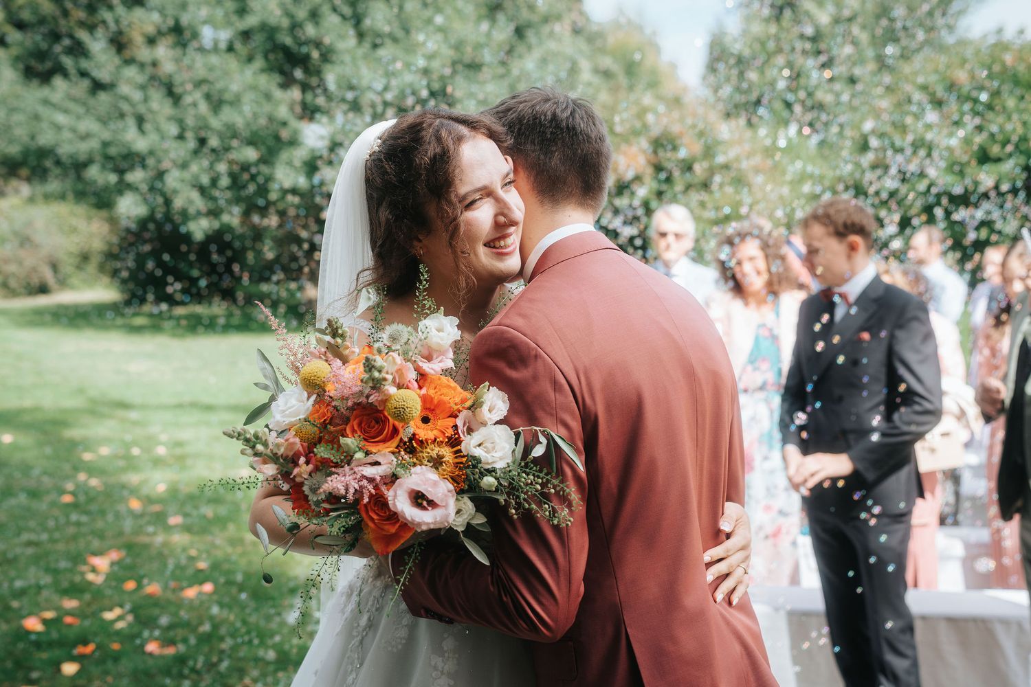 Smiling bride in a veil holds an orange and white bouquet while embracing a groom in a rust suit as wedding guests watch in a sunny garden.