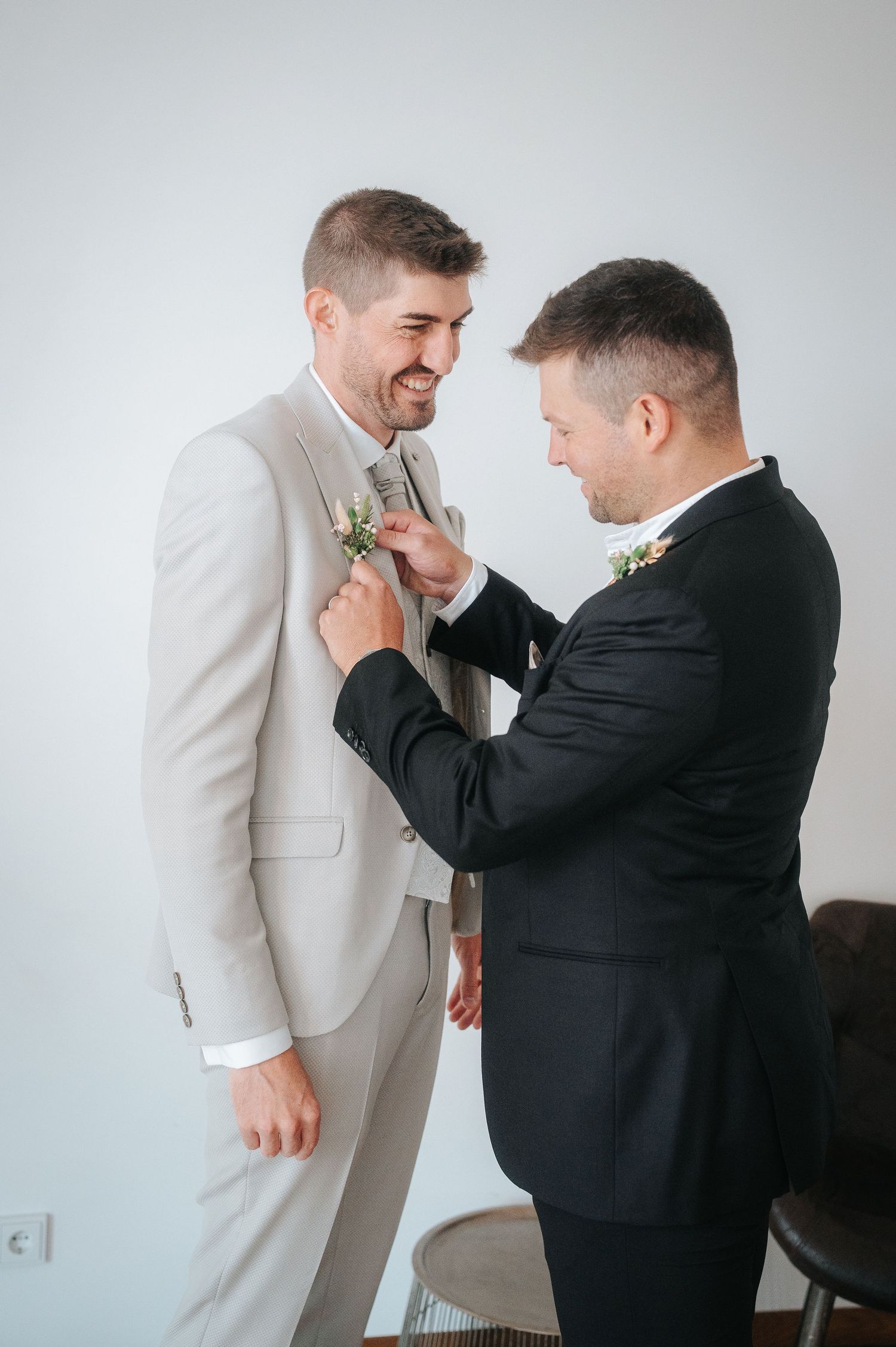 Two men in wedding attire, one in a beige suit smiling while the other in a black suit pins a floral boutonniere onto his lapel.