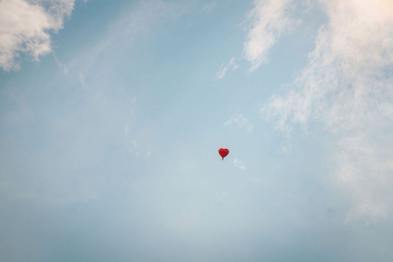 A small red heart-shaped balloon floats alone in a soft blue sky with wispy white clouds.