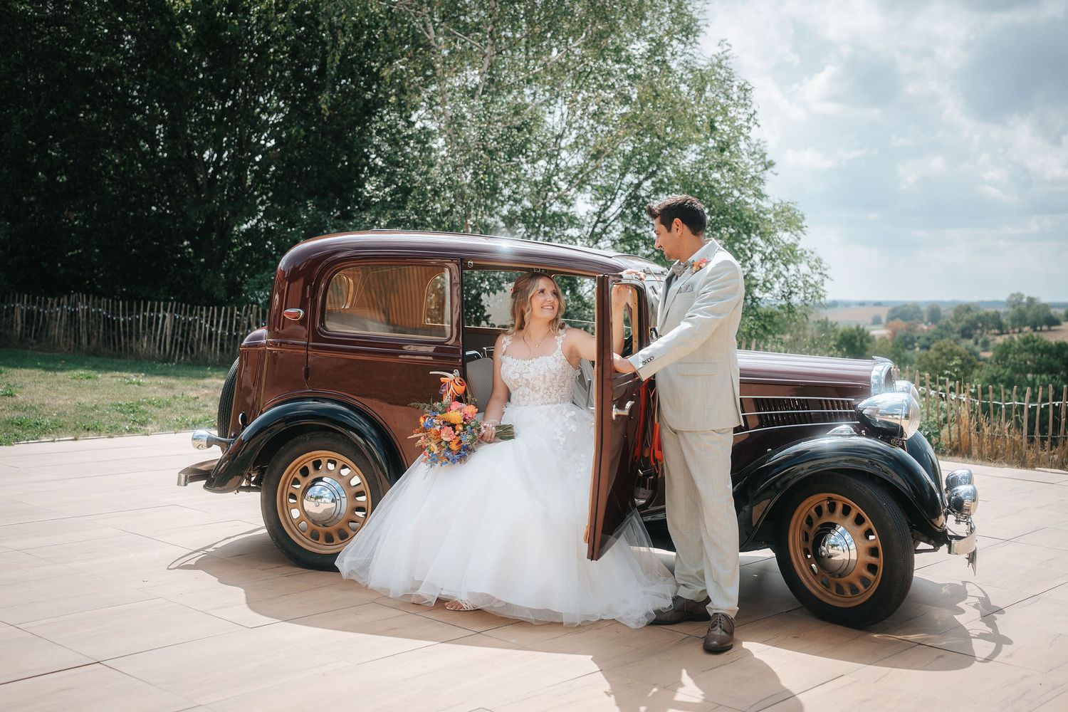 Bride in a white wedding gown holding an orange bouquet sits in a vintage brown car as the groom in a gray suit opens the door.