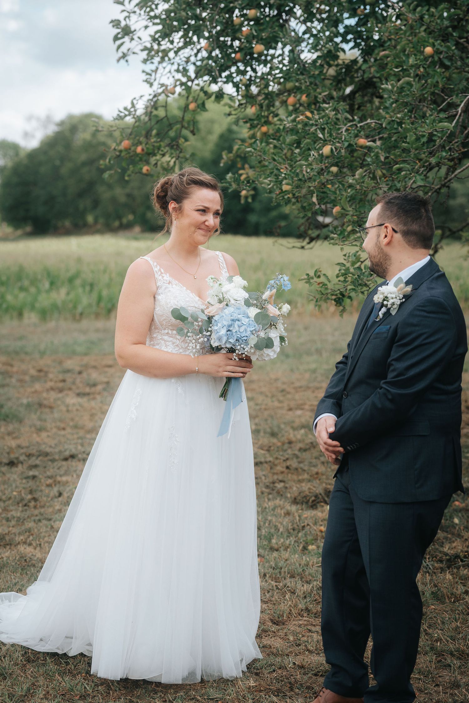 Smiling bride in a white lace gown holds a blue and white bouquet while facing the groom in a navy suit beneath an apple tree.