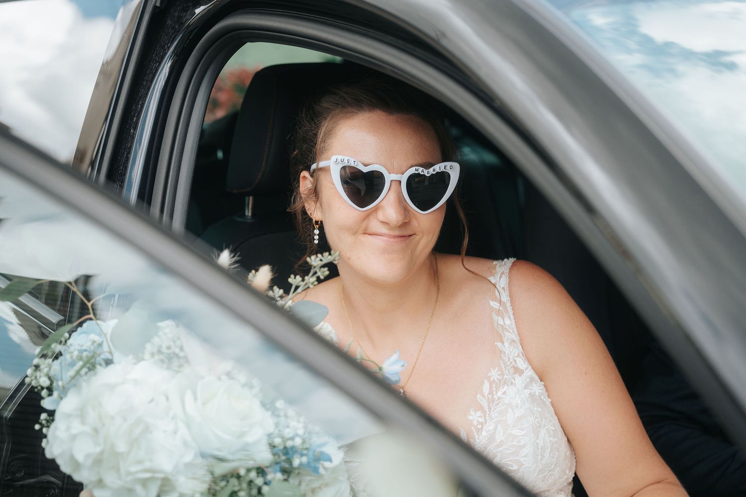 Smiling bride in a lace wedding dress and heart-shaped sunglasses looks out a car window beside a white floral bouquet.