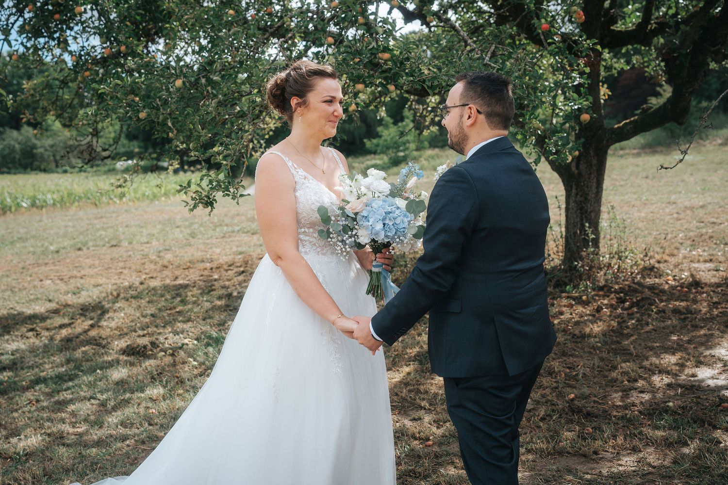 Bride in a white lace gown holding a blue and white bouquet joins hands with a groom in a dark suit beneath an orchard tree.