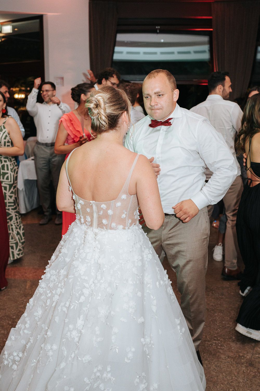 Two people dancing at a wedding reception, with the bride wearing a white lace gown with sheer back detailing.