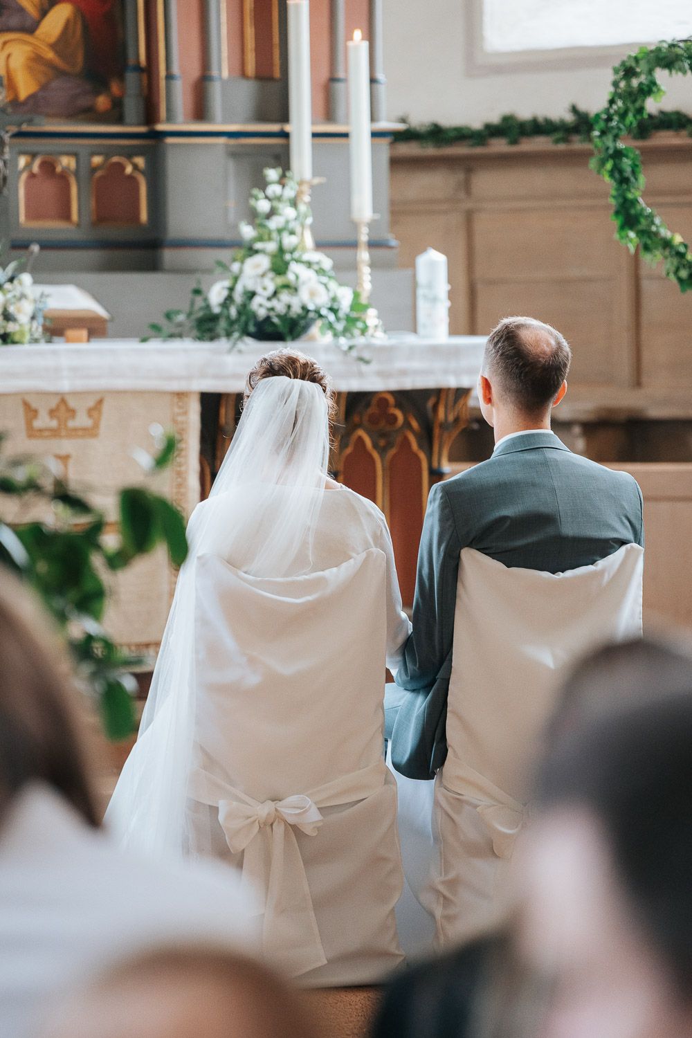 A couple sits together during their traditional church wedding ceremony with white bow-adorned chair covers.