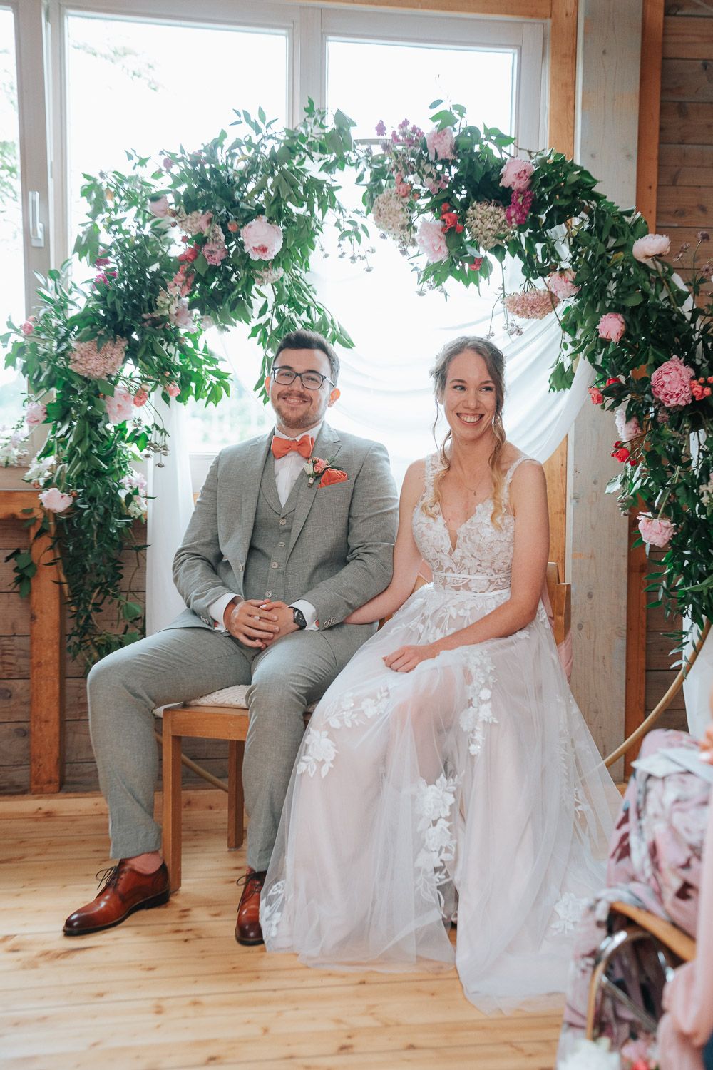 A couple sits together in a rustic indoor setting decorated with pink floral arrangements and greenery.