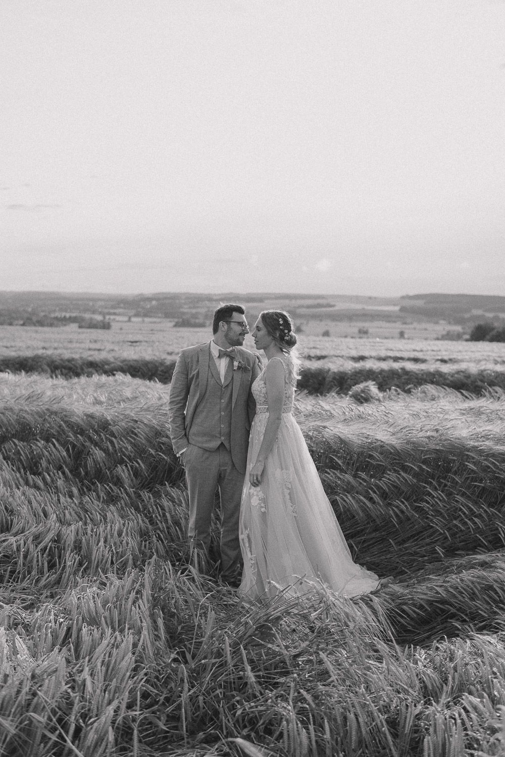 A black and white wedding photo showing a couple standing in a windswept field of tall grass with rolling hills in the background.