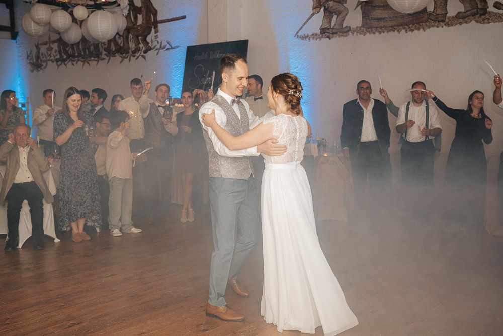 A couple shares their first dance as newlyweds on a misty dance floor with guests watching in the background.