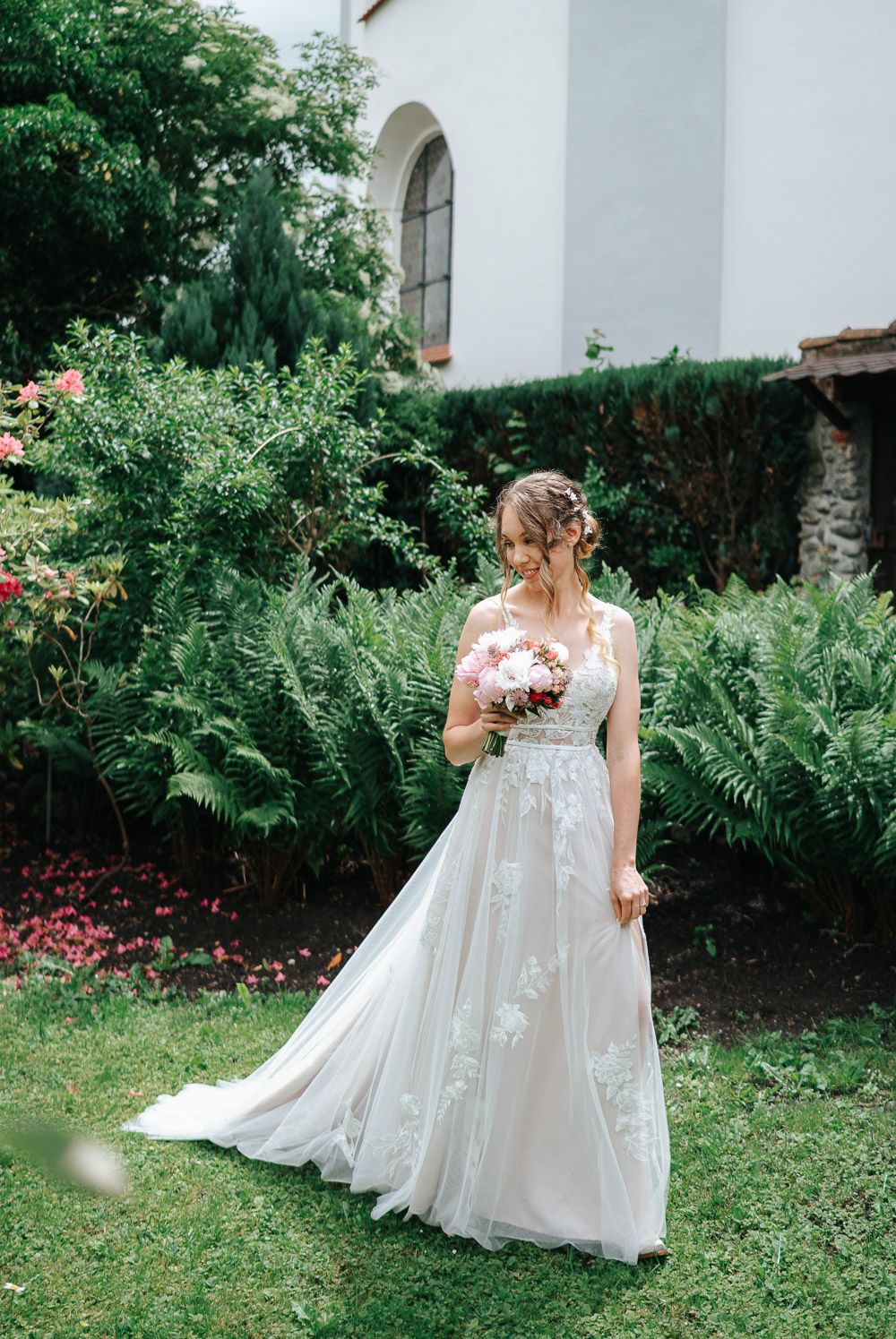 A bride in a flowing lace wedding gown stands in a garden with ferns and arched church windows in the background.