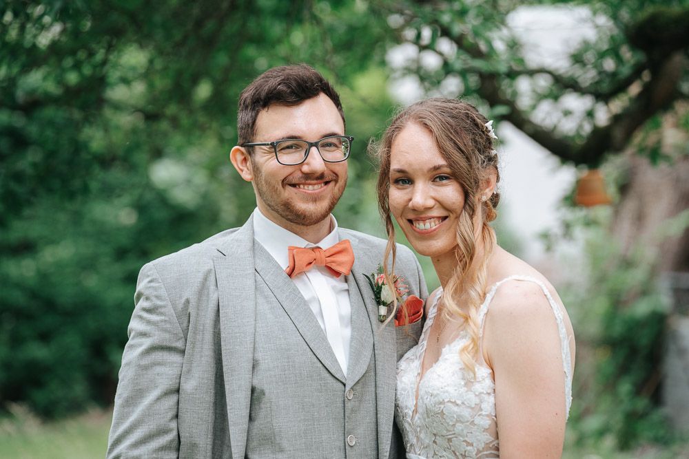 A bride and groom smile together outdoors, with the groom wearing a gray suit and coral bow tie, surrounded by greenery.