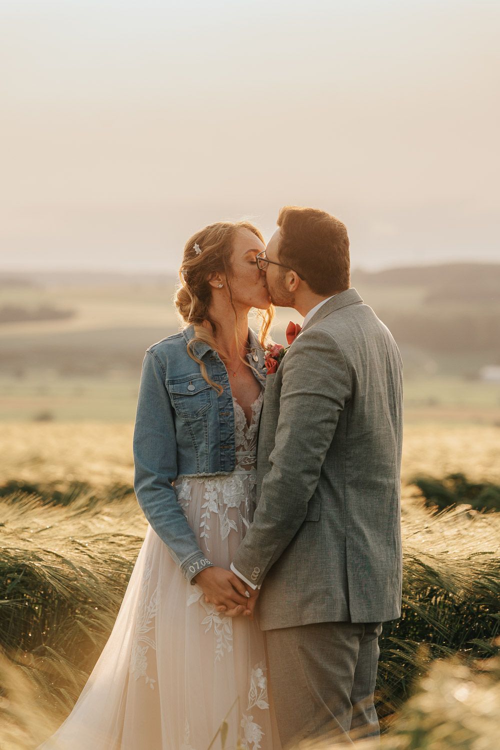 A couple shares a romantic kiss in a wheat field at sunset, bride wearing a white dress and denim jacket.
