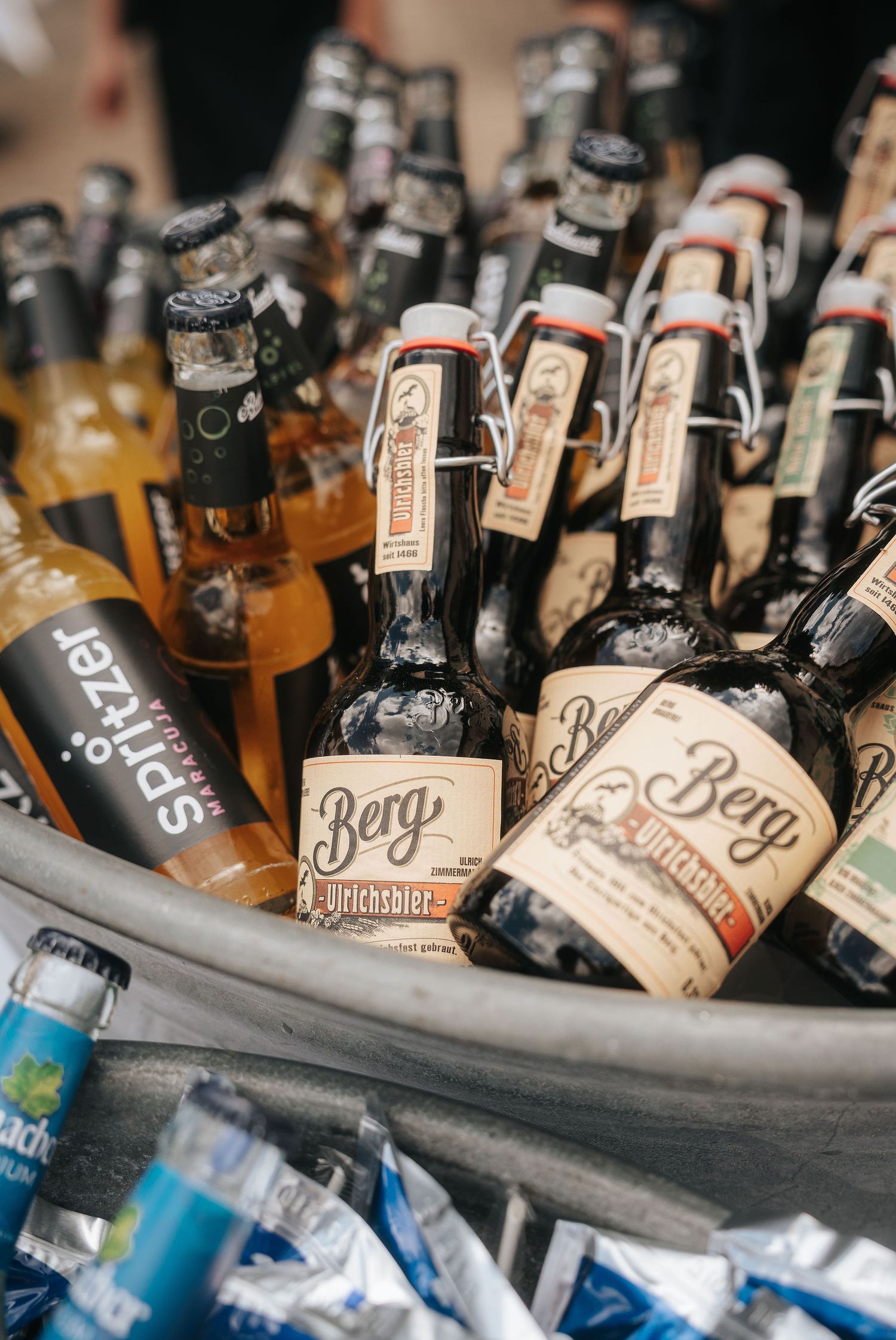 Berg beer bottles with swing-top caps chilling in a metal tub of ice alongside Spritzer bottles and blue drink cans.