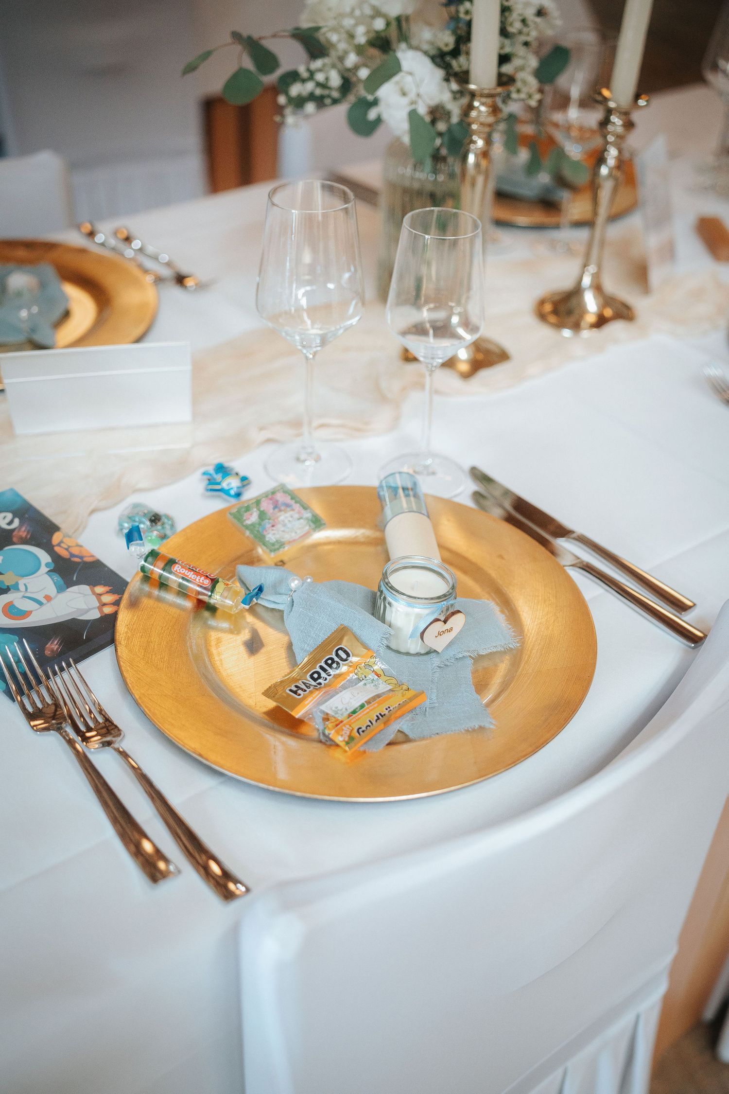 Elegant wedding table setting with a gold charger plate, wine glasses, rose gold cutlery, candles, and small favors on a white tablecloth.