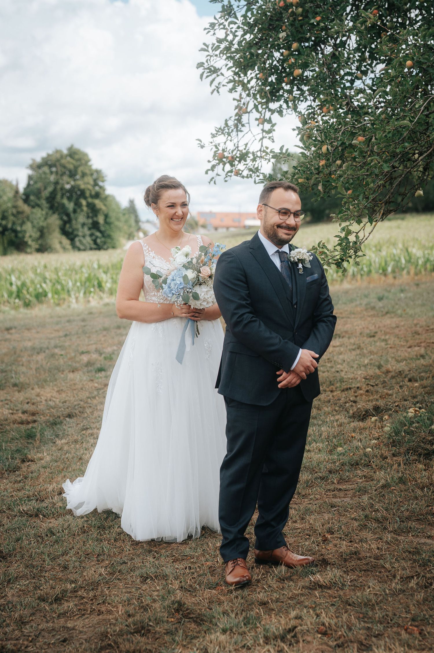 Smiling bride in a white tulle gown holding a blue bouquet stands behind a groom in a navy suit under an apple tree in a sunny field.