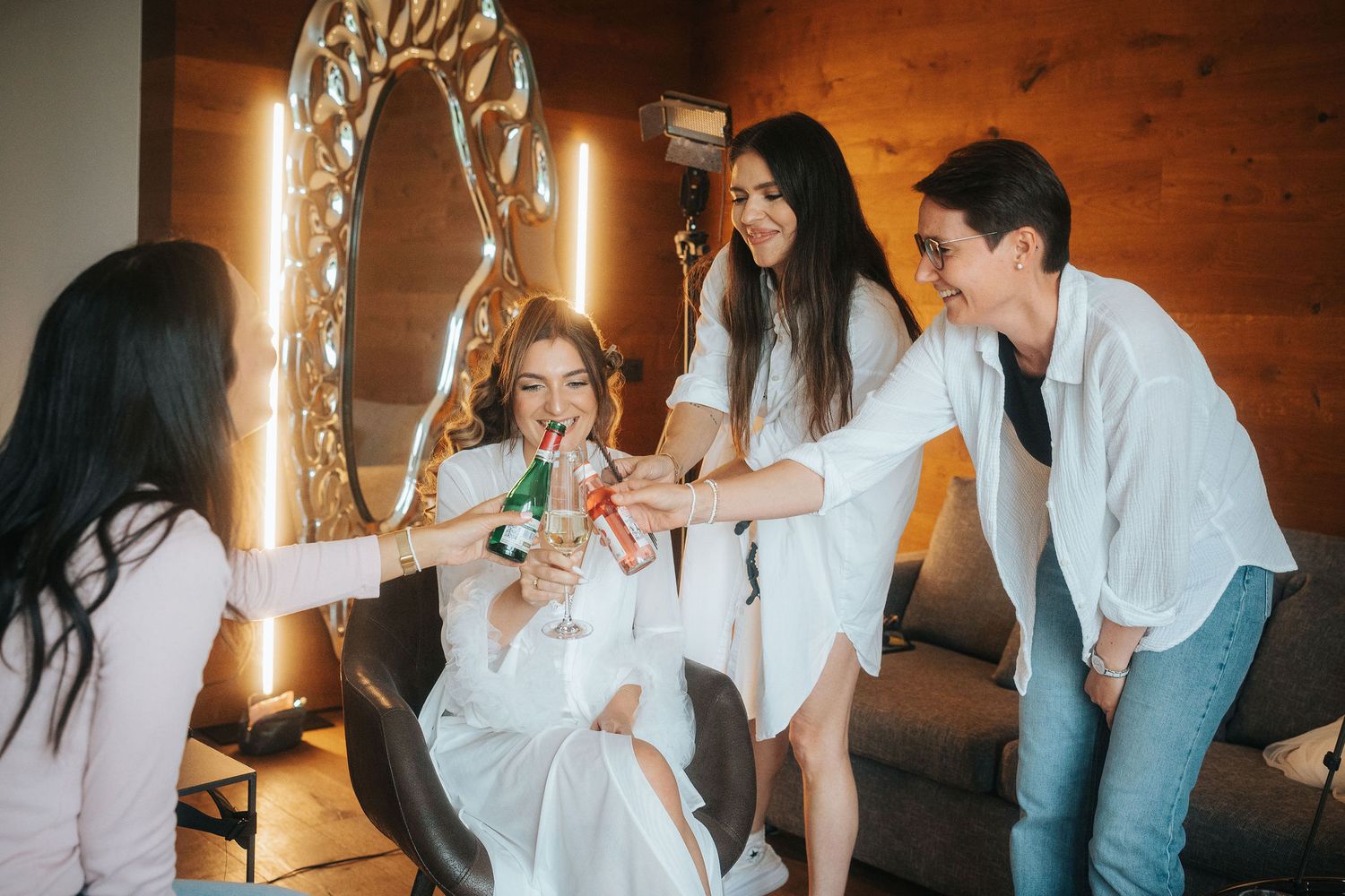 Four smiling women in white outfits clink colorful drinks together in a cozy room with a lit vanity mirror and wooden walls.