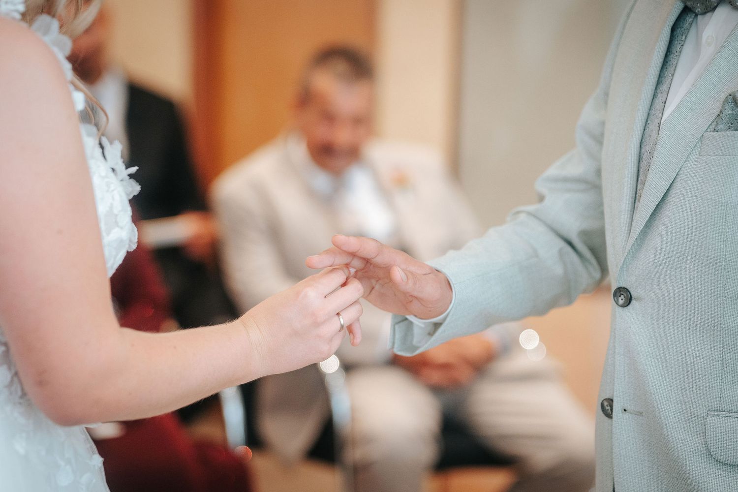 Bride in a lace dress and groom in a light gray suit exchange wedding rings during a ceremony as guests look on.