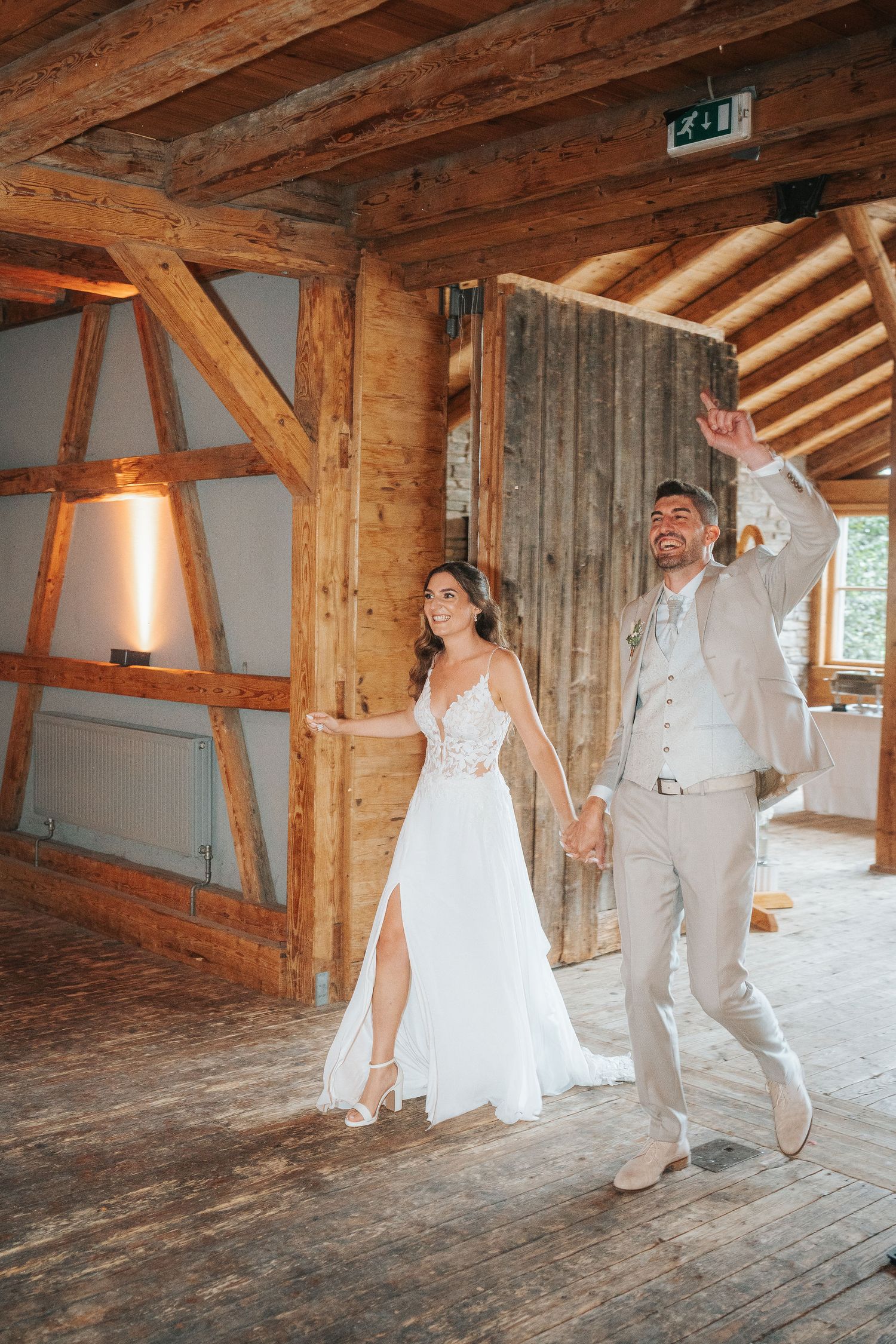 Smiling bride in a white lace gown and groom in a beige suit walk hand in hand through a rustic wooden barn wedding venue.