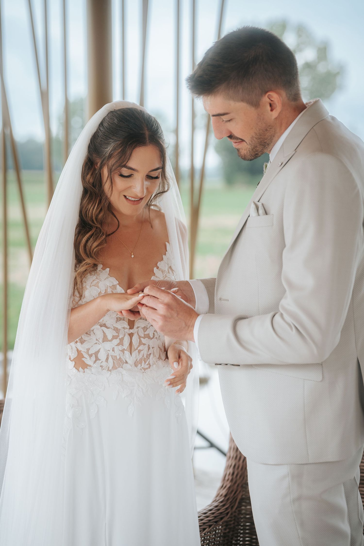 Smiling bride in a lace wedding gown and veil exchanges rings with the groom in a light beige suit during an outdoor ceremony.