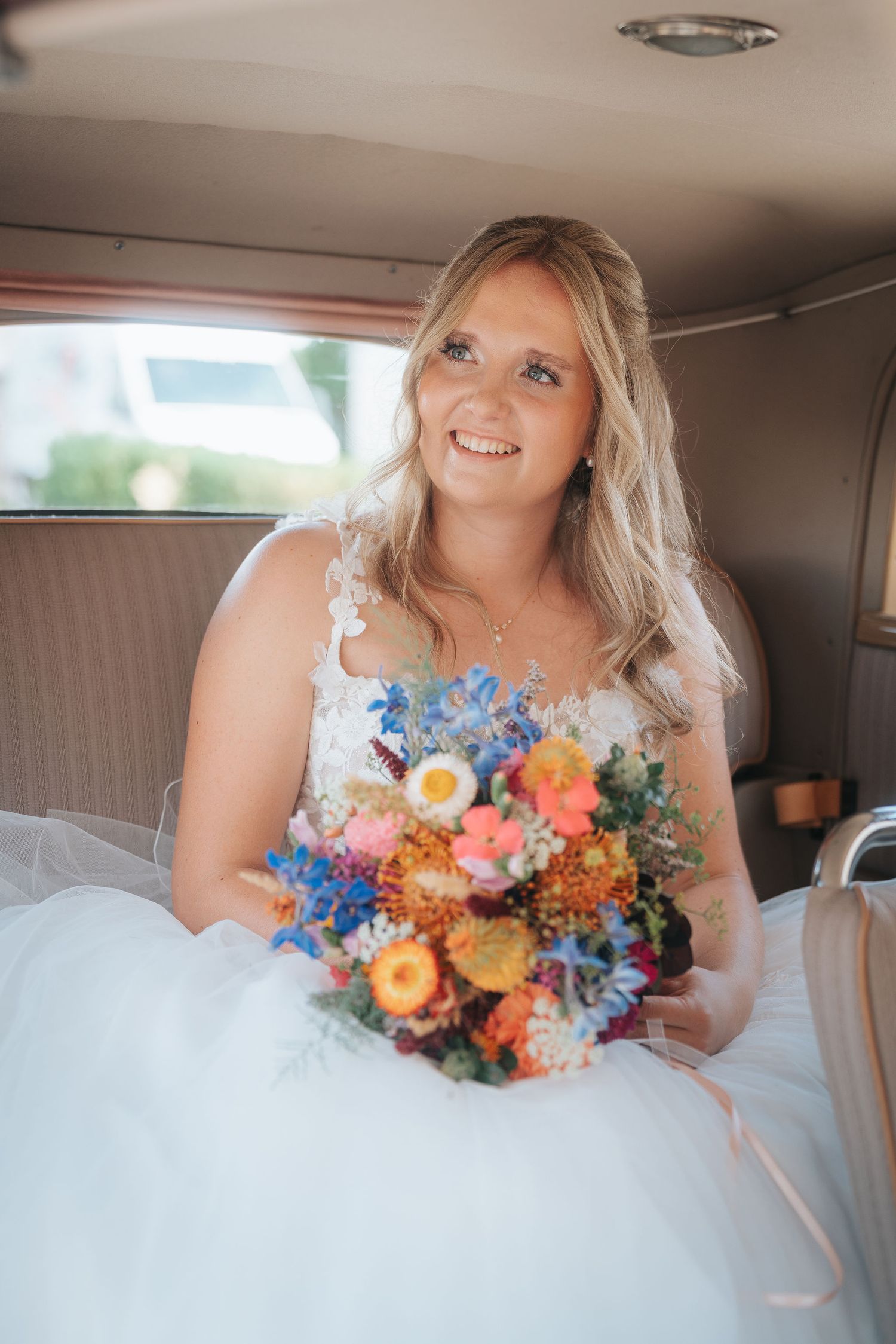 Smiling bride in a white lace wedding gown holds a colorful wildflower bouquet while seated in the back of a vintage car.