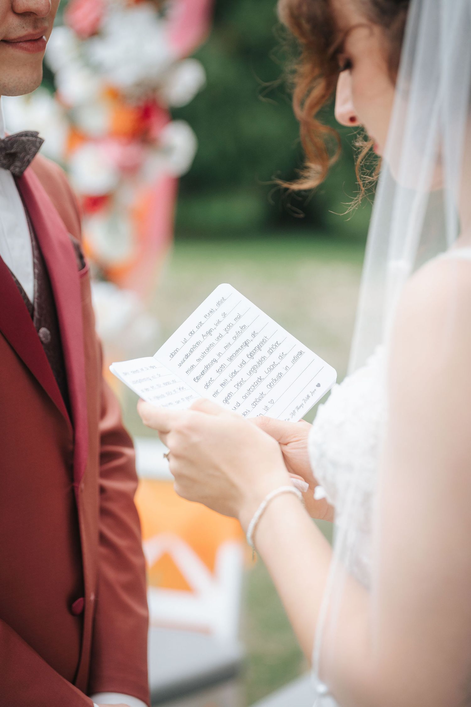 Bride in a white lace dress and veil reads handwritten vows from a notebook to a groom in a burgundy suit at an outdoor wedding.