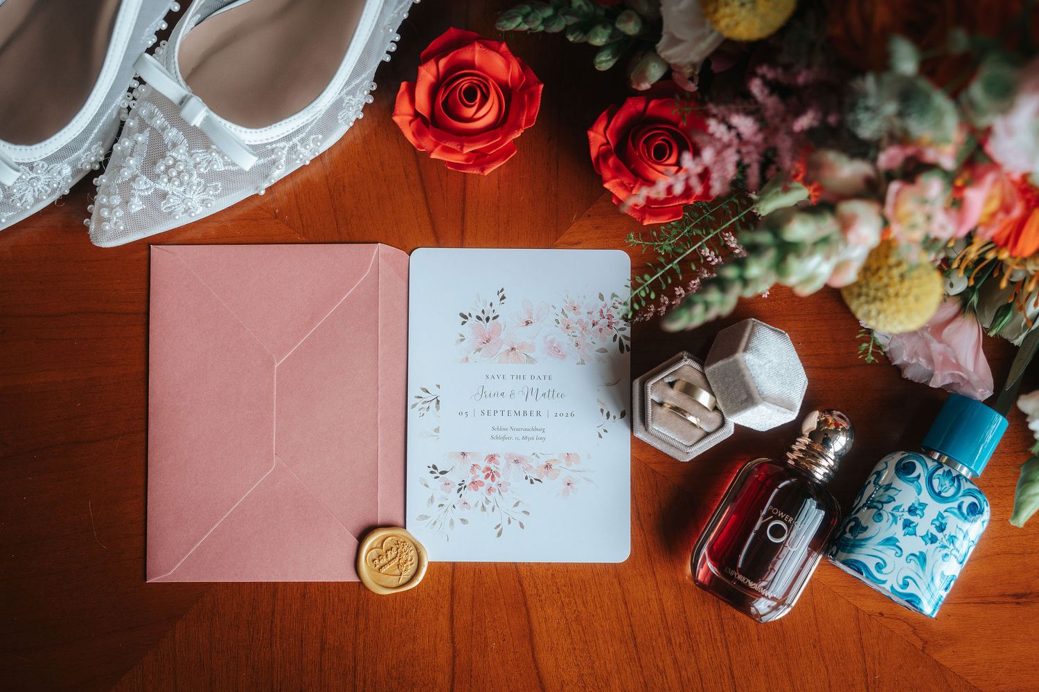 Wedding flat lay with a pink envelope, floral invitation, wax seal, ring box, perfume bottles, bridal heels and bouquet on a wooden table.