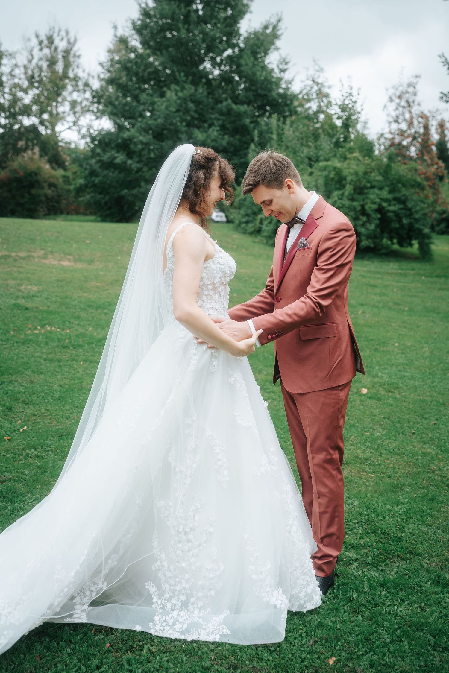 Bride in a lace wedding gown with a long veil holds hands with a groom in a rust-colored suit on a green lawn surrounded by trees.