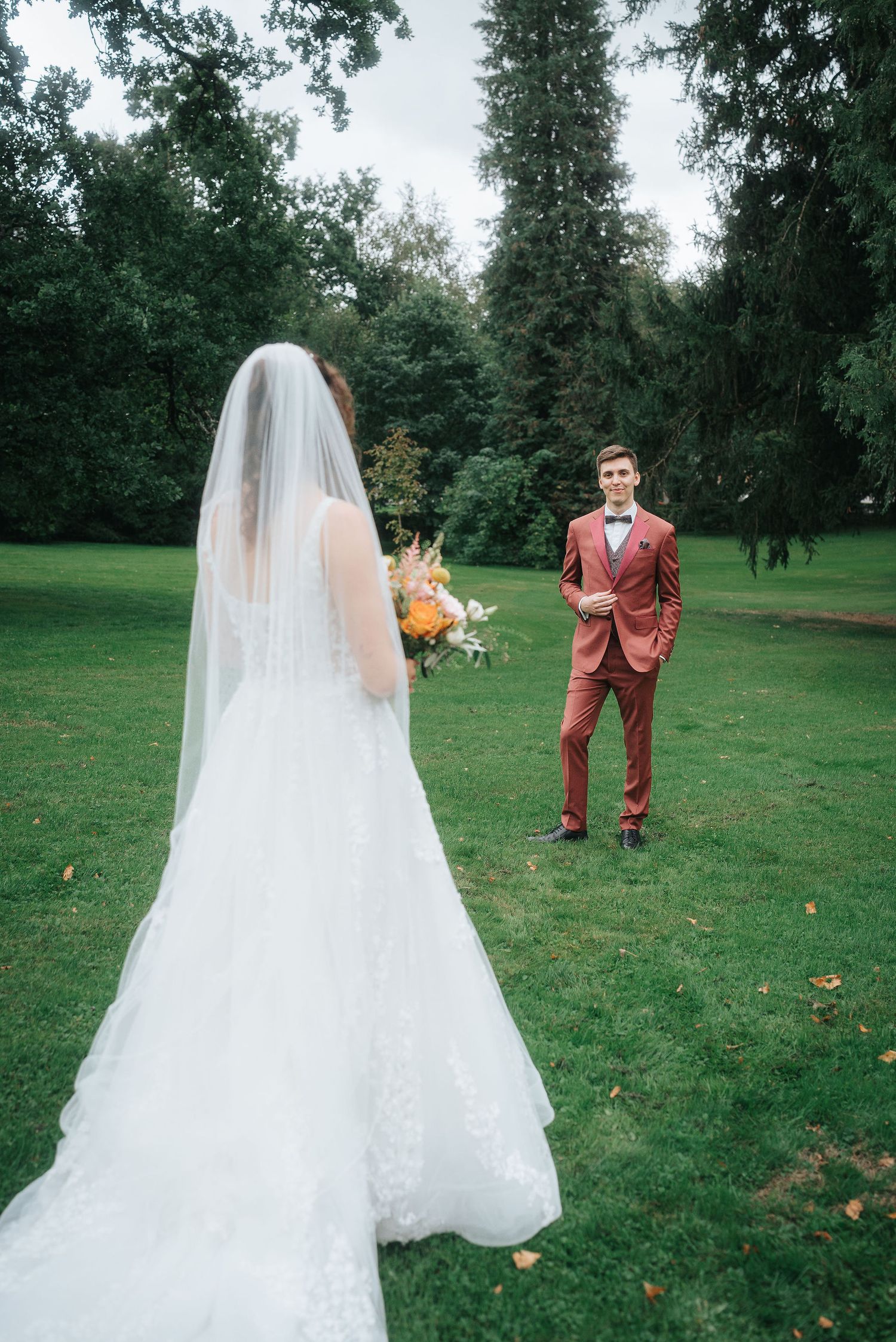 Bride in a lace gown and long veil holds an orange bouquet while facing a groom in a rust suit on a green lawn surrounded by tall trees.