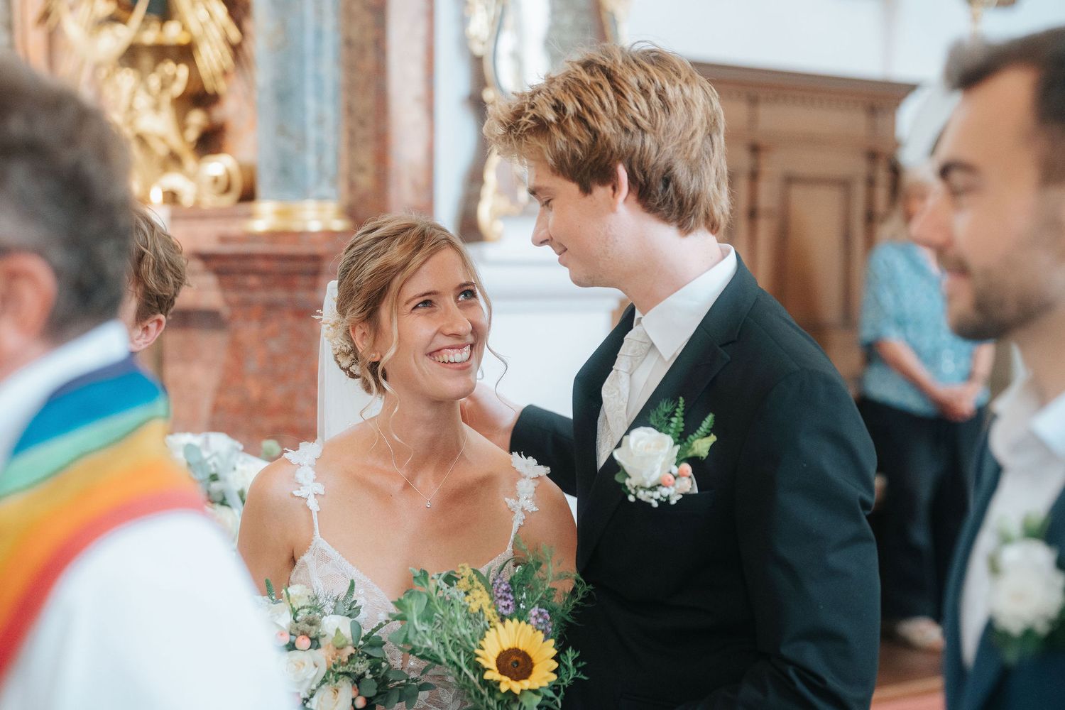 Smiling bride holding a sunflower bouquet shares a joyful moment with a groom in a dark suit during a wedding ceremony in a church.
